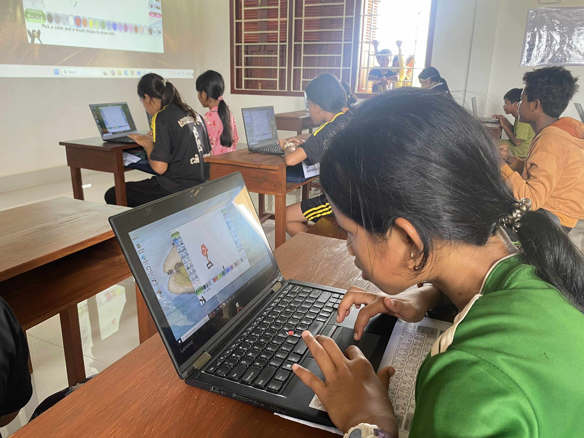 Students in a classroom, using laptops. A projector displays on the wall.