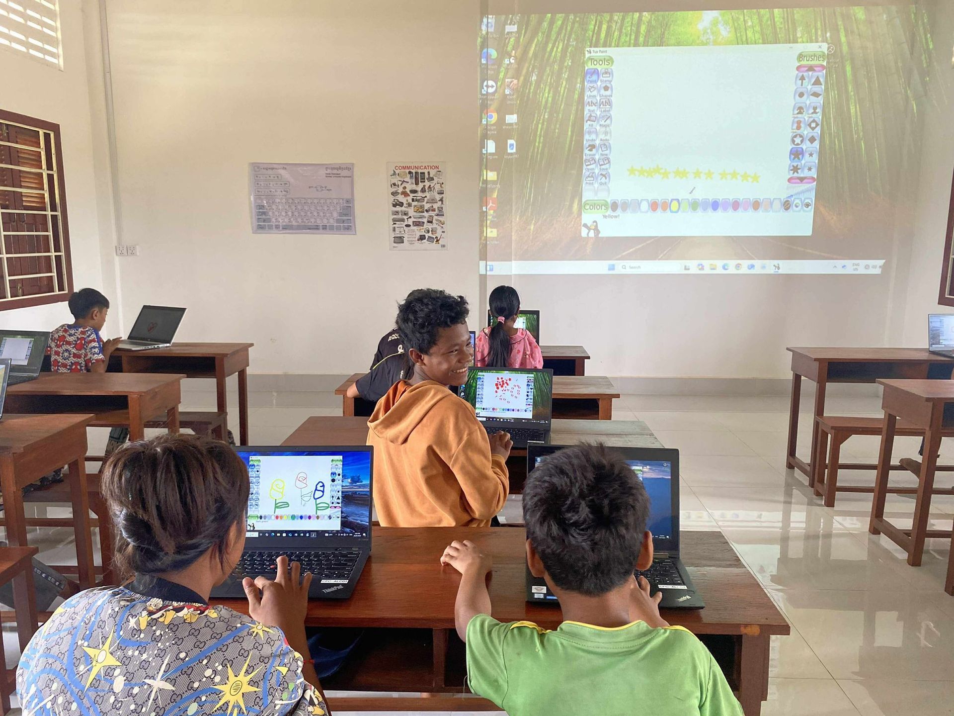 Children in a computer lab, looking at laptops, some facing the camera. Projector screen displaying an image.