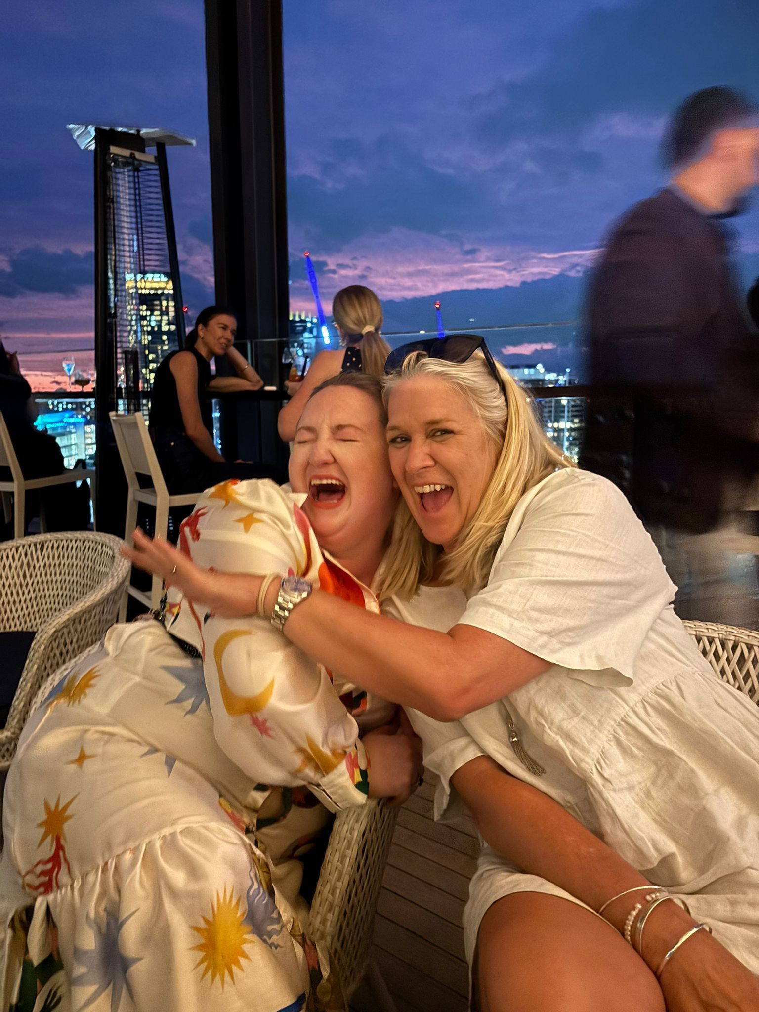 Two women laughing and hugging on an outdoor patio at dusk; city skyline in background.