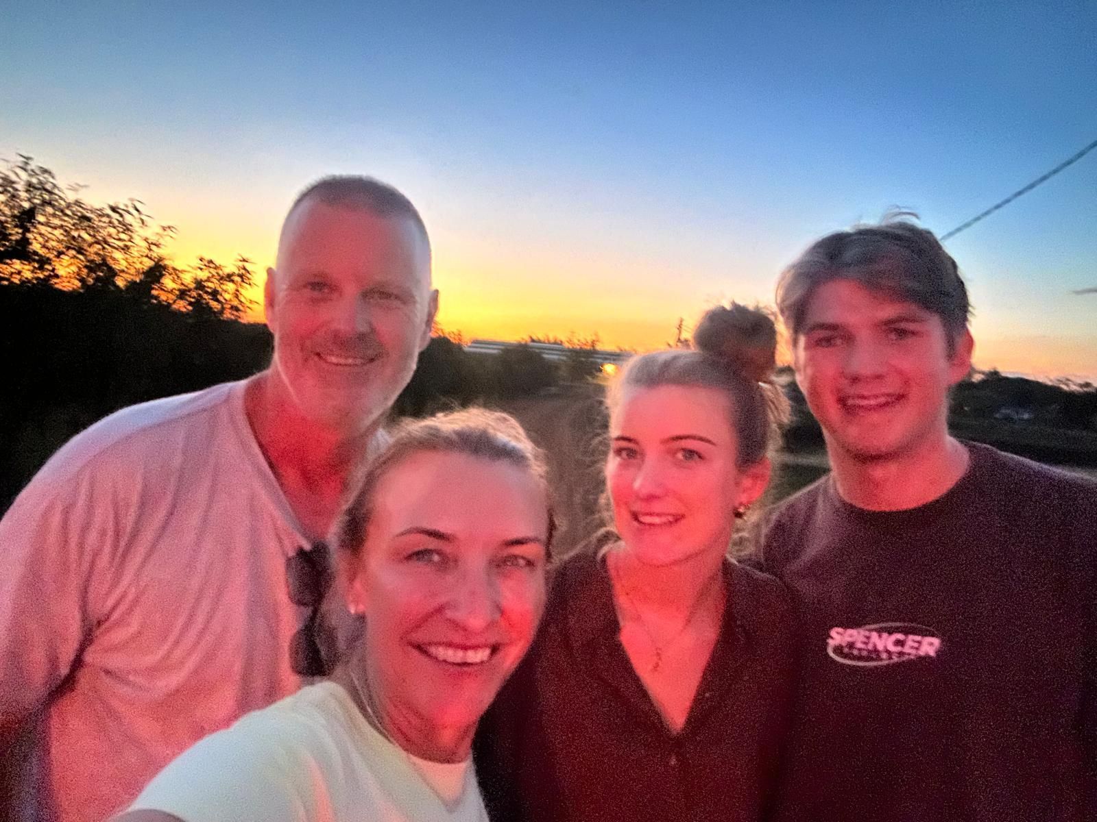 Family selfie at sunset; sky orange and blue, four smiling people on a road.