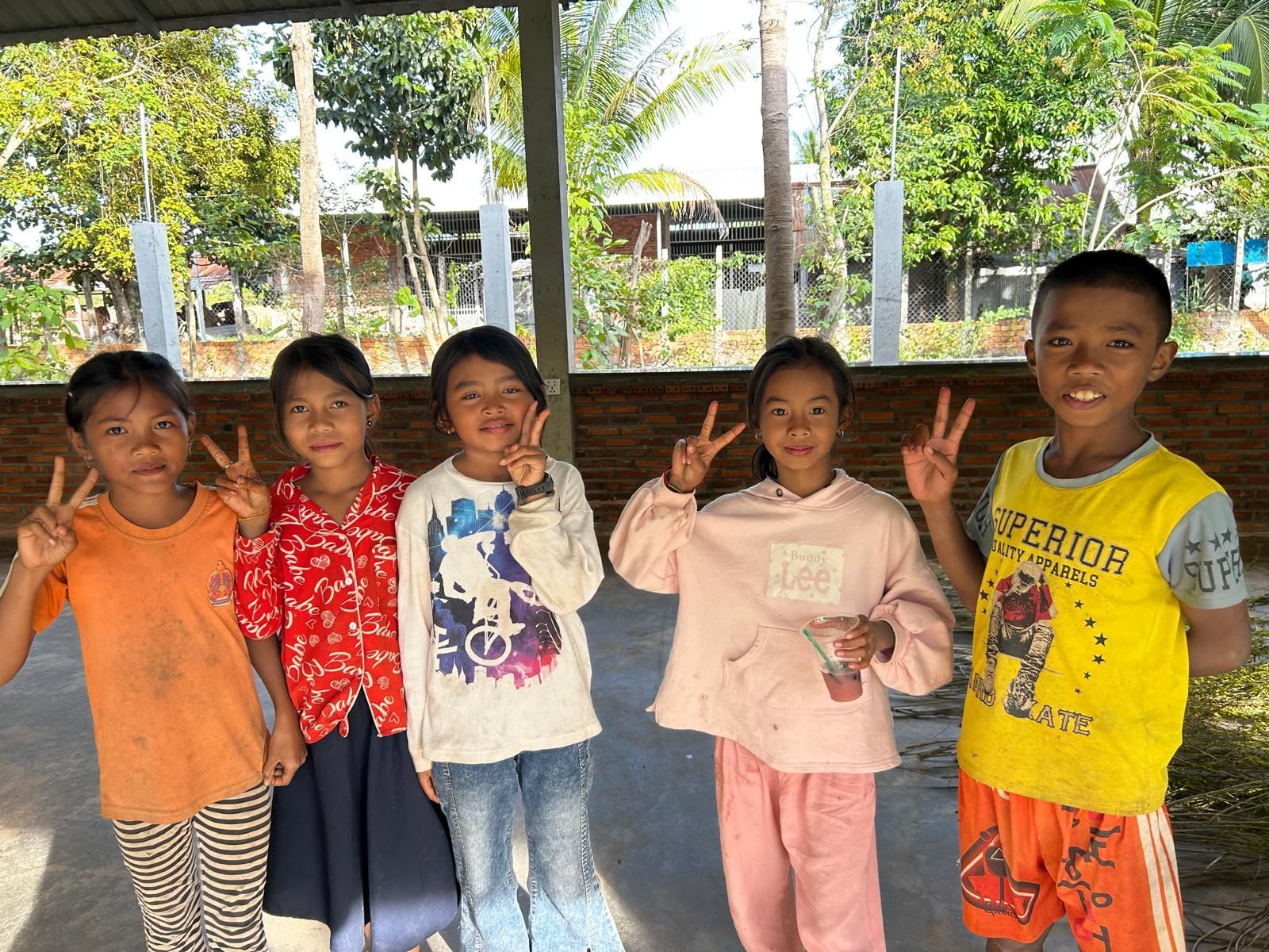 Five children smiling and giving peace signs outdoors. Sunlight, trees, and a wooden structure are visible.