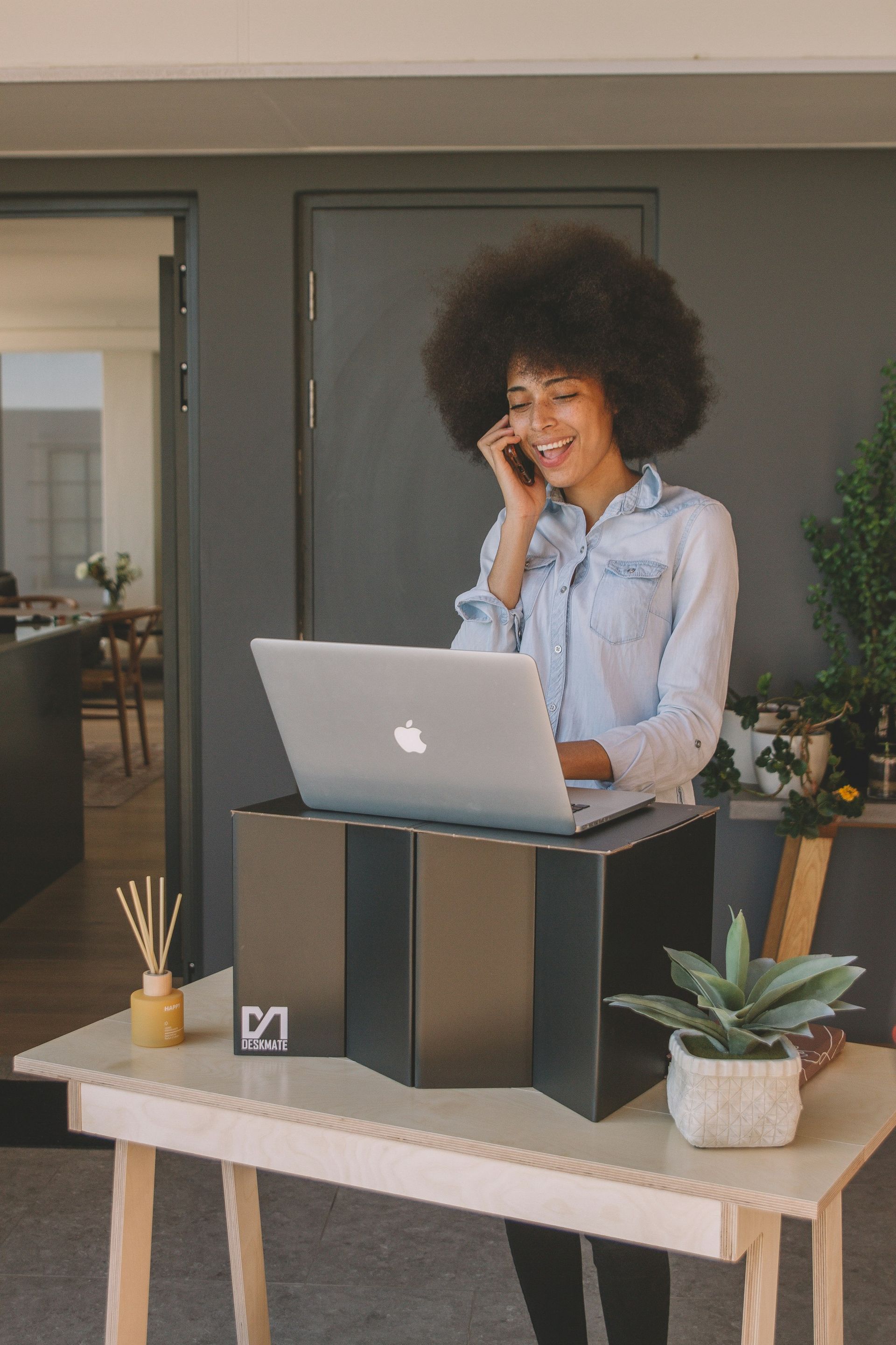 Woman on the phone, using a laptop on a standing desk made of cardboard, in a room with plants.