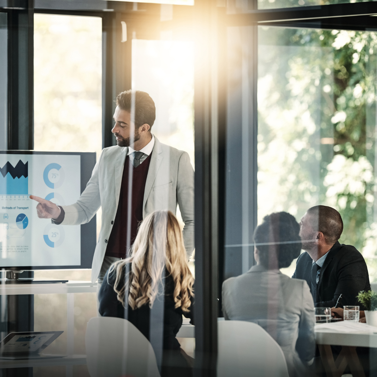 Man in suit presenting data on a screen to a team in a modern office with natural light.