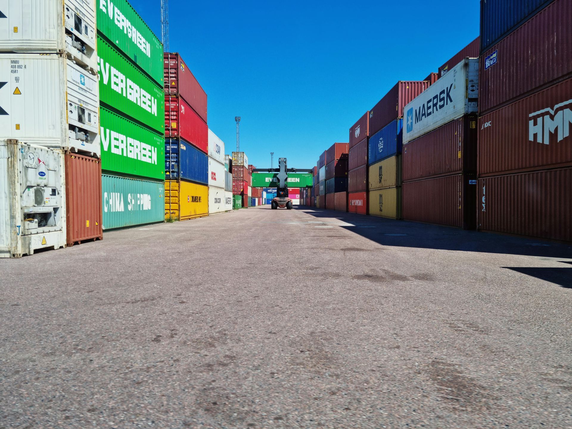 A shipping container yard with stacks of colorful containers on either side of a gravel path under a clear blue sky.