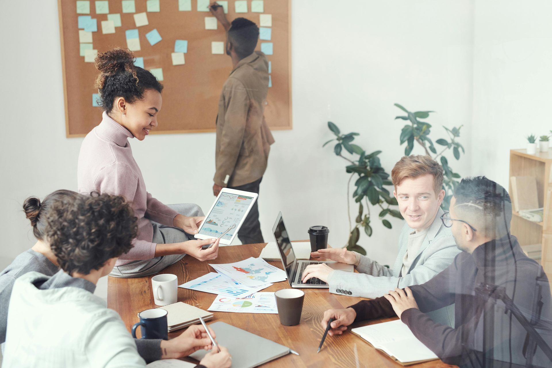 People in a meeting, some working on laptops and papers, others by a corkboard with sticky notes.