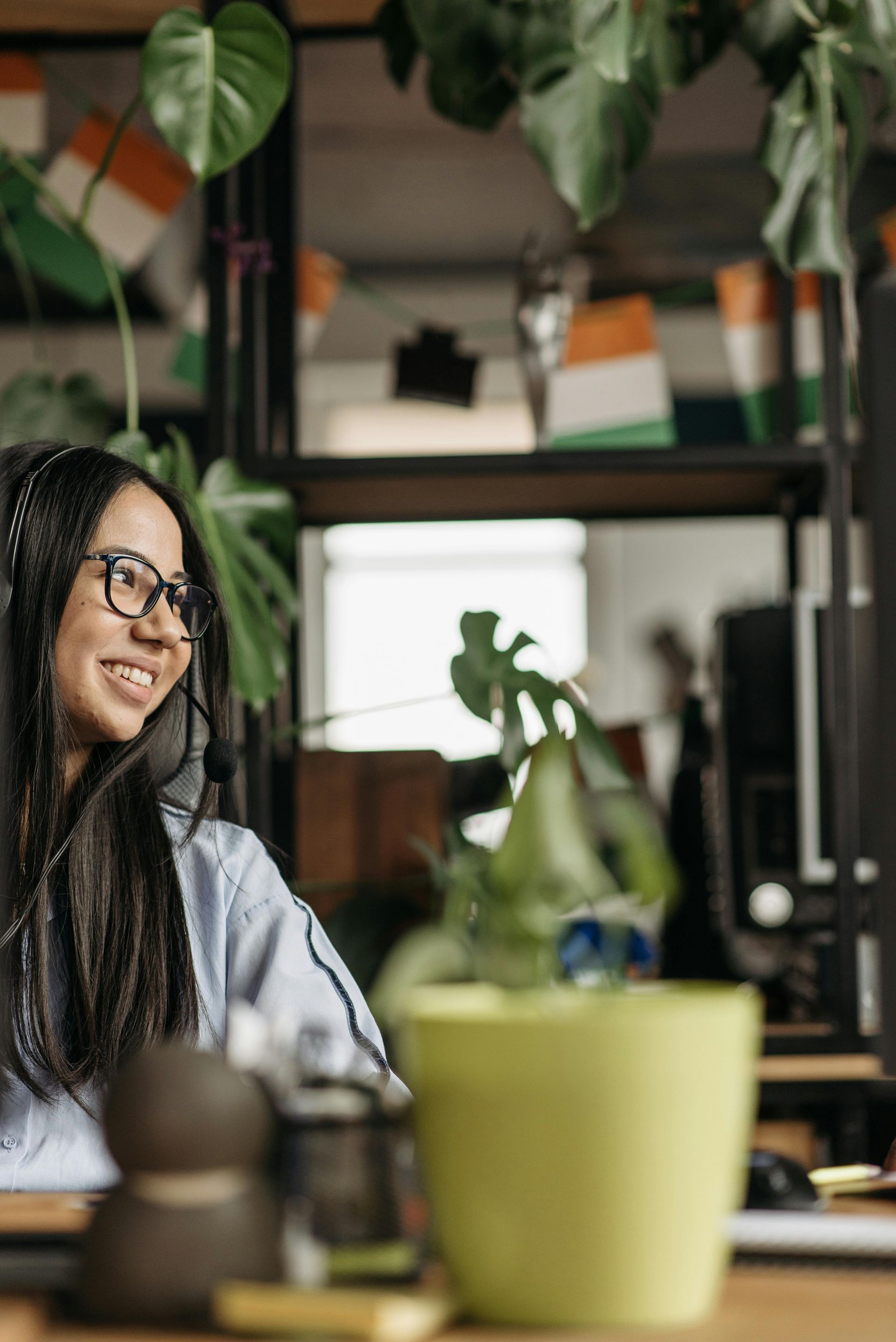 Woman with glasses smiling, wearing headset, working at desk with plants in office.