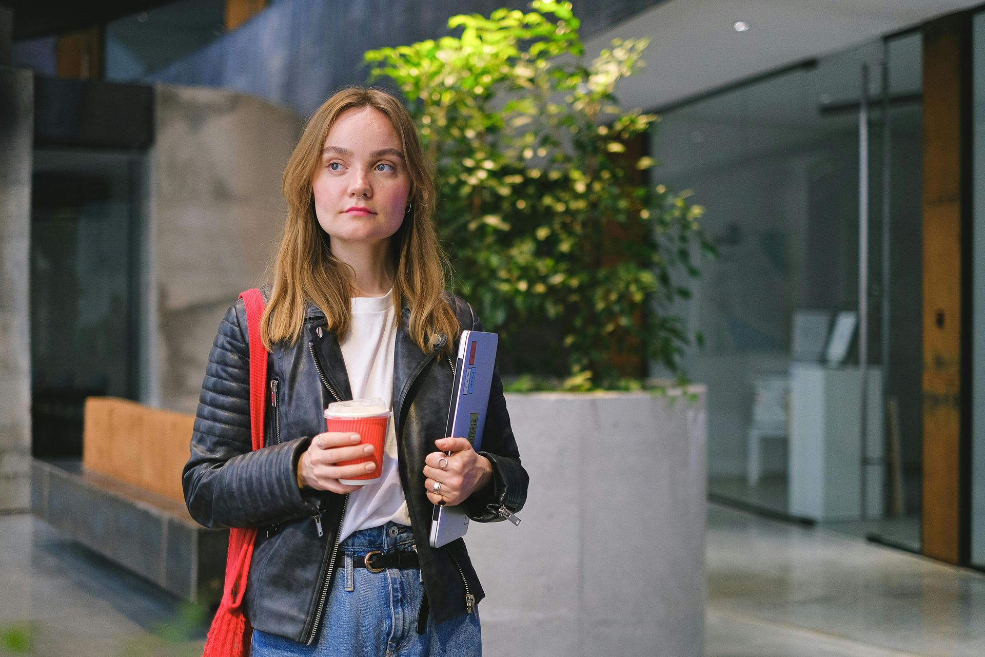 Woman in leather jacket, holding coffee and laptop, in modern building with greenery.