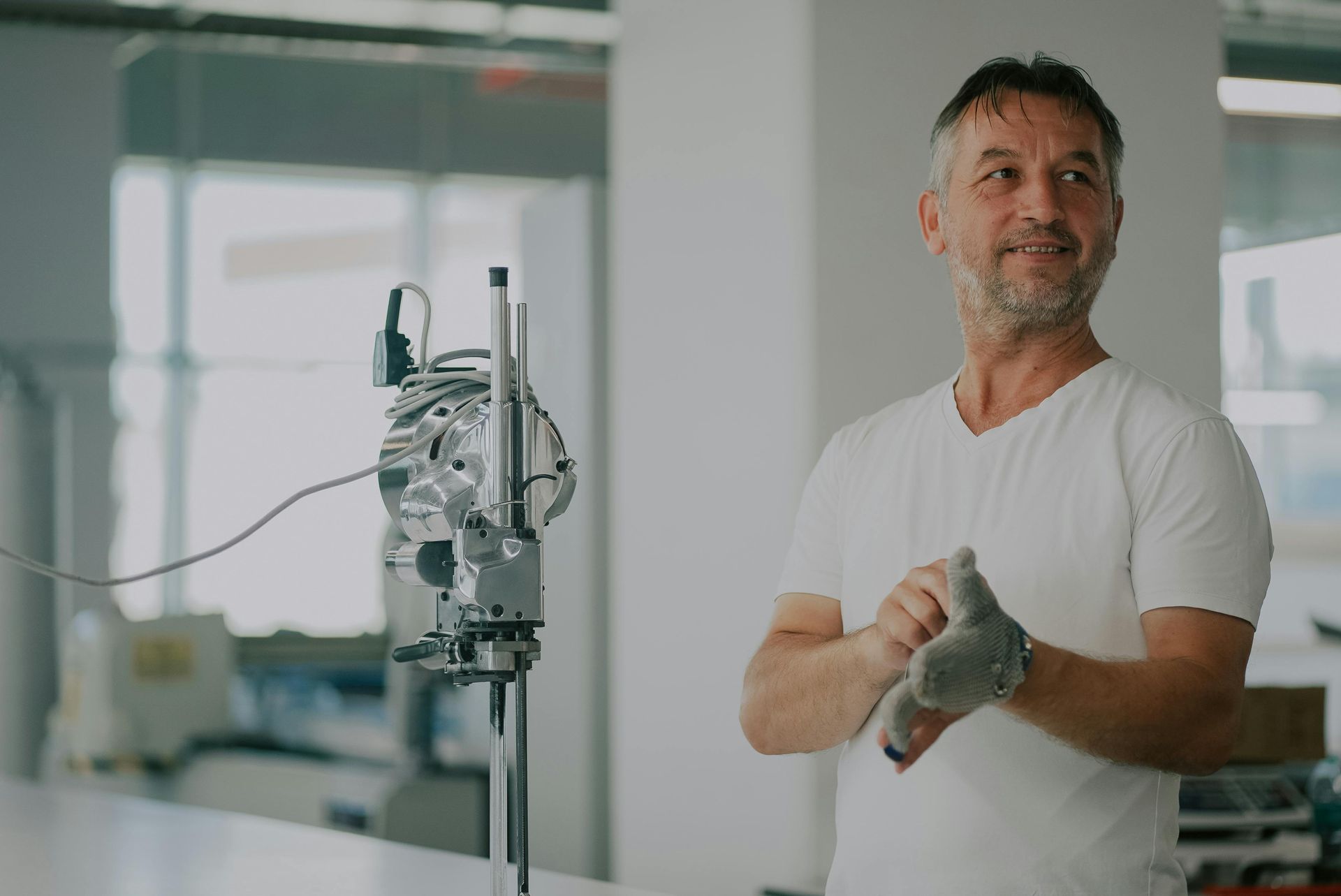 Man in a white shirt putting on a glove, smiling, in a bright workshop with machinery.