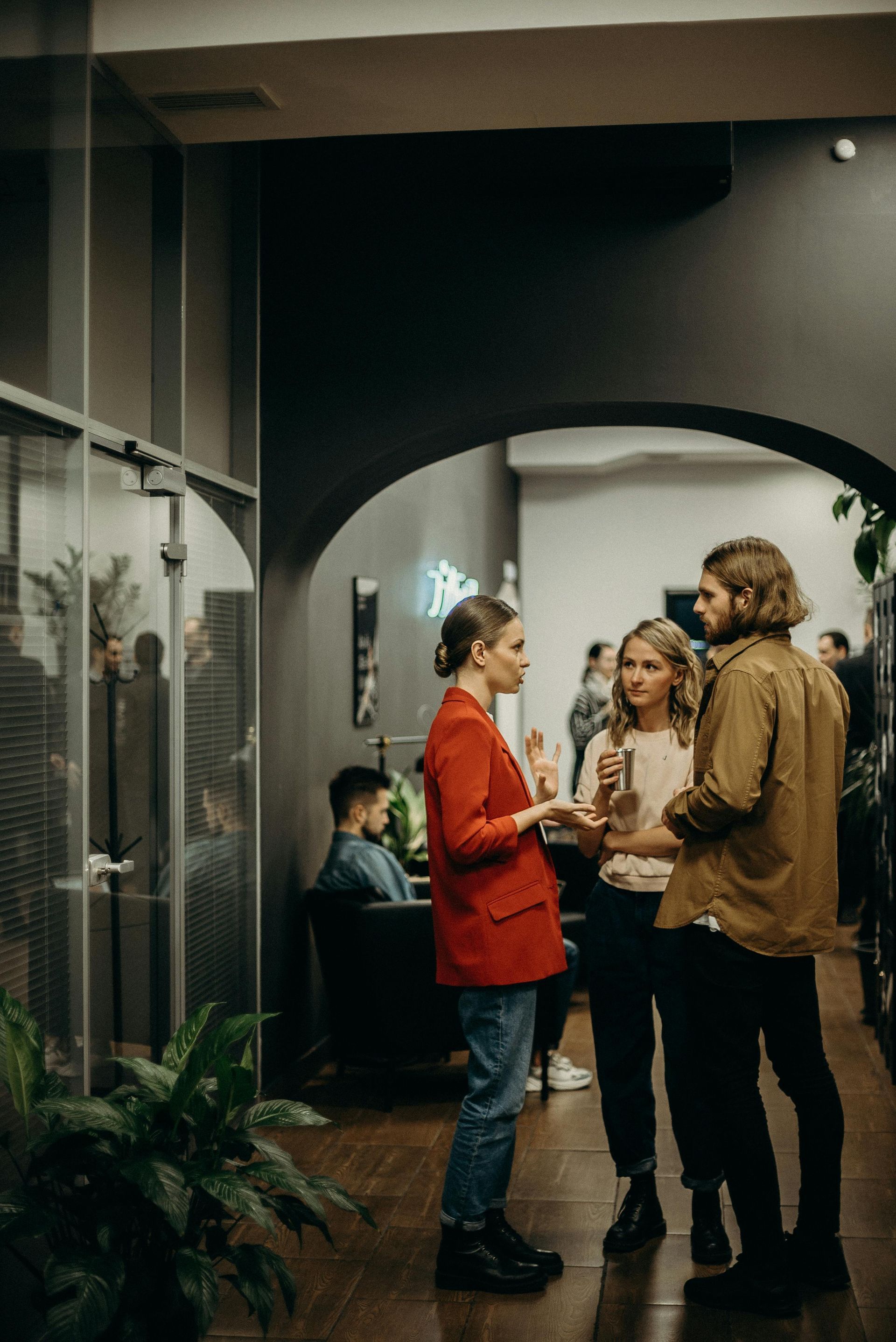 Three people conversing in a dimly lit hallway; one in a red blazer, plants on the left, others in the background.