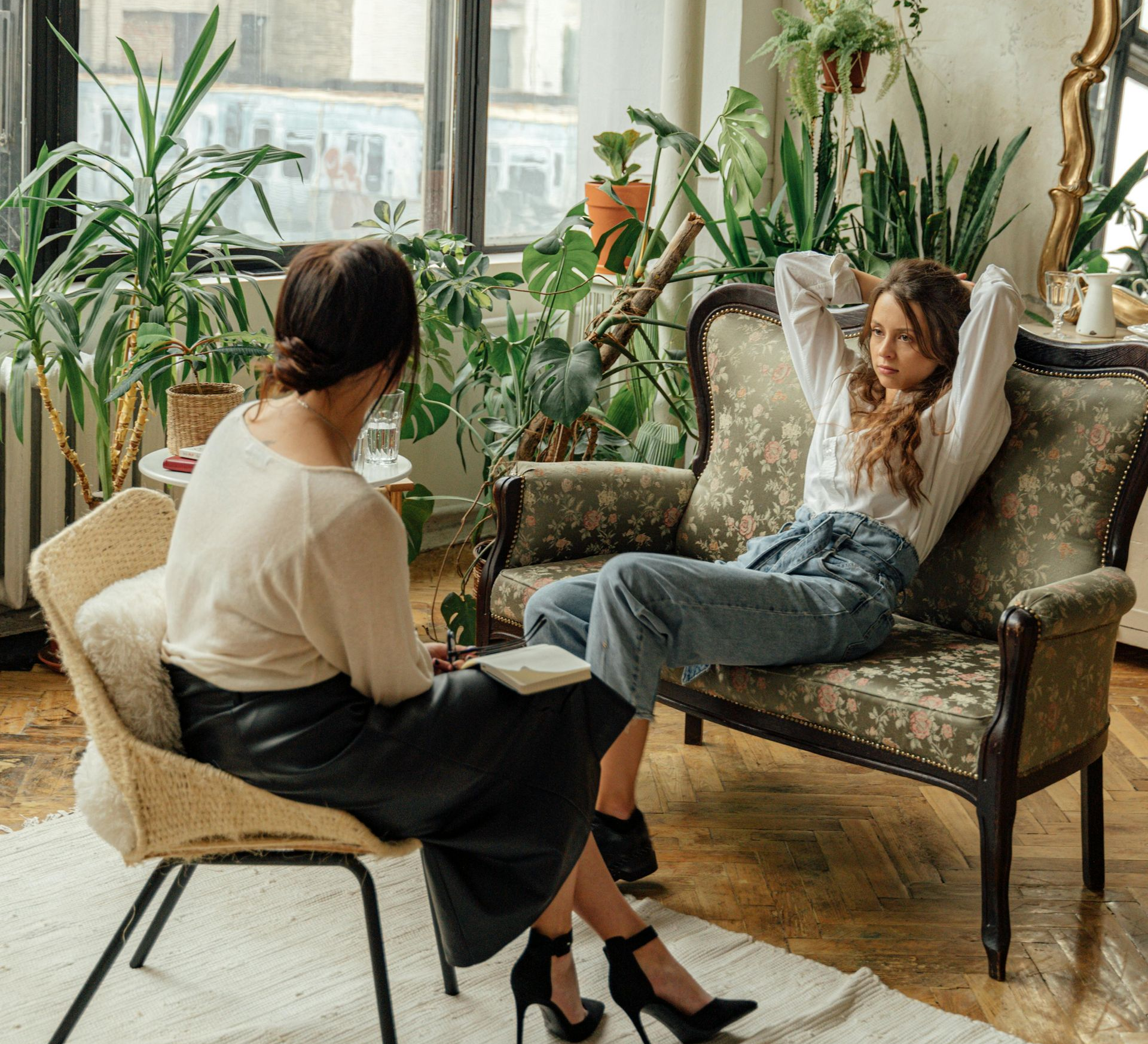 A woman consults with a therapist in a room with many plants. The woman sits on a couch, arms behind her head.
