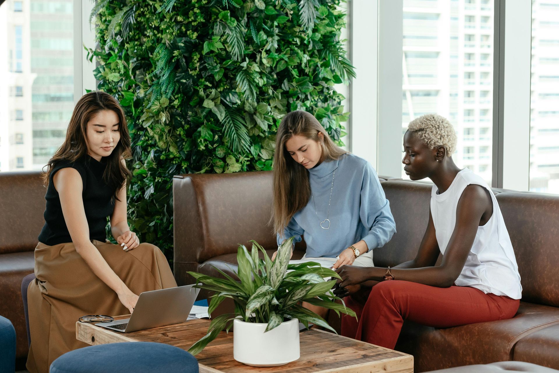 Three people collaborating on couch, one using laptop, greenery in background.