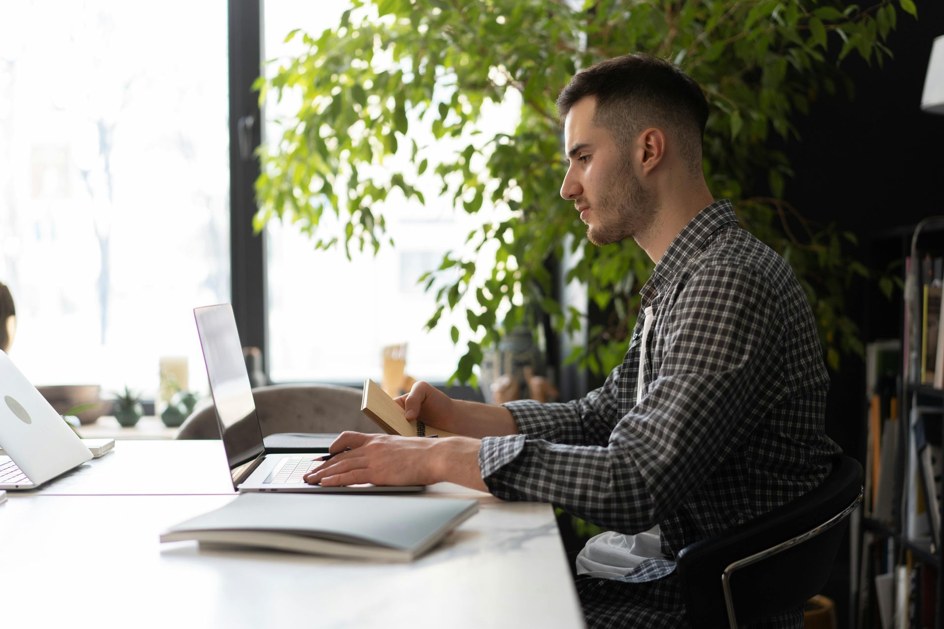 Man working on a laptop at a table, holding a small book. Bright window and plants in the background.