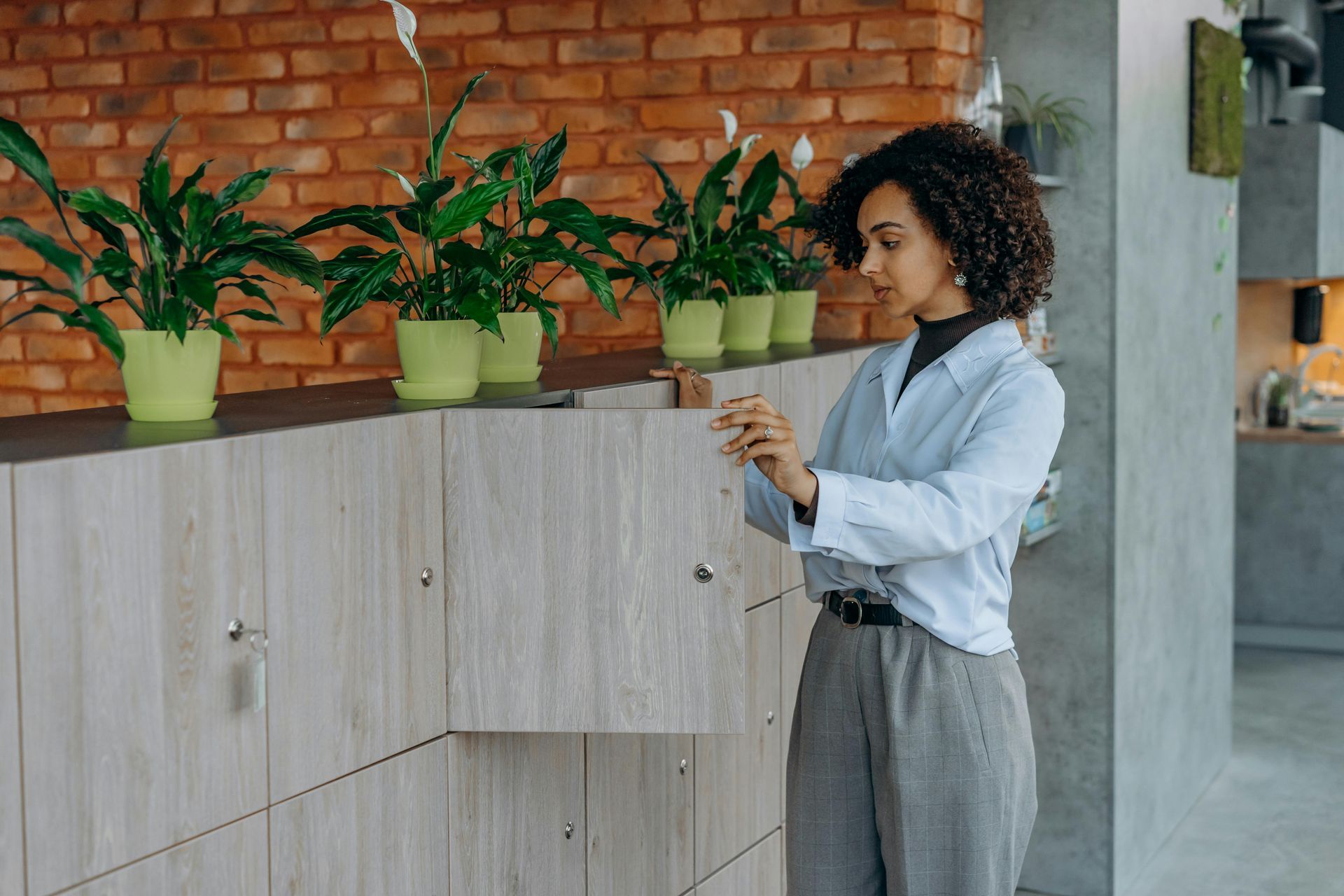 Woman in a light blue shirt tending potted plants on a wooden shelf against a brick wall.
