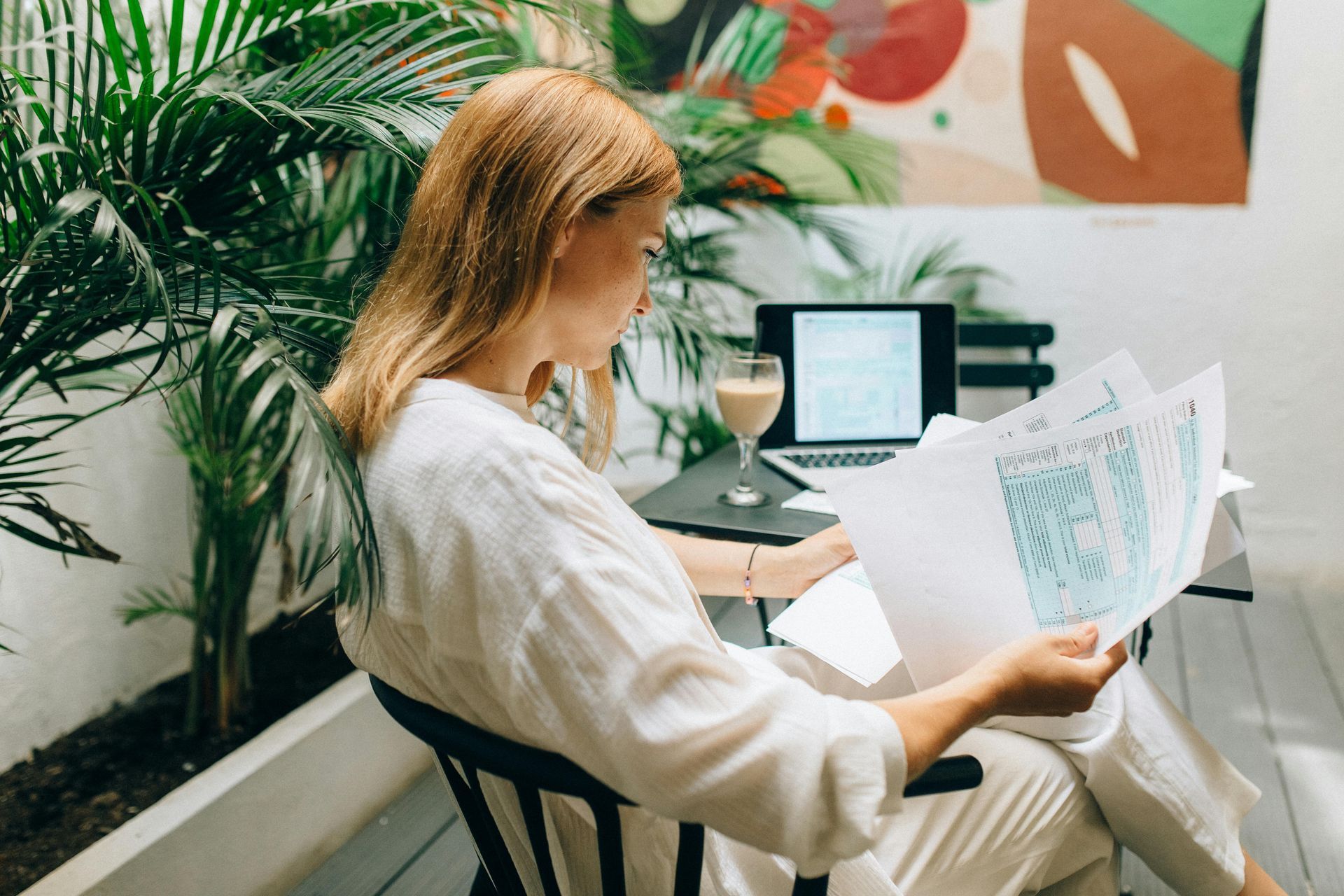 Woman in white, looking at papers, laptop on table with drink, surrounded by plants.