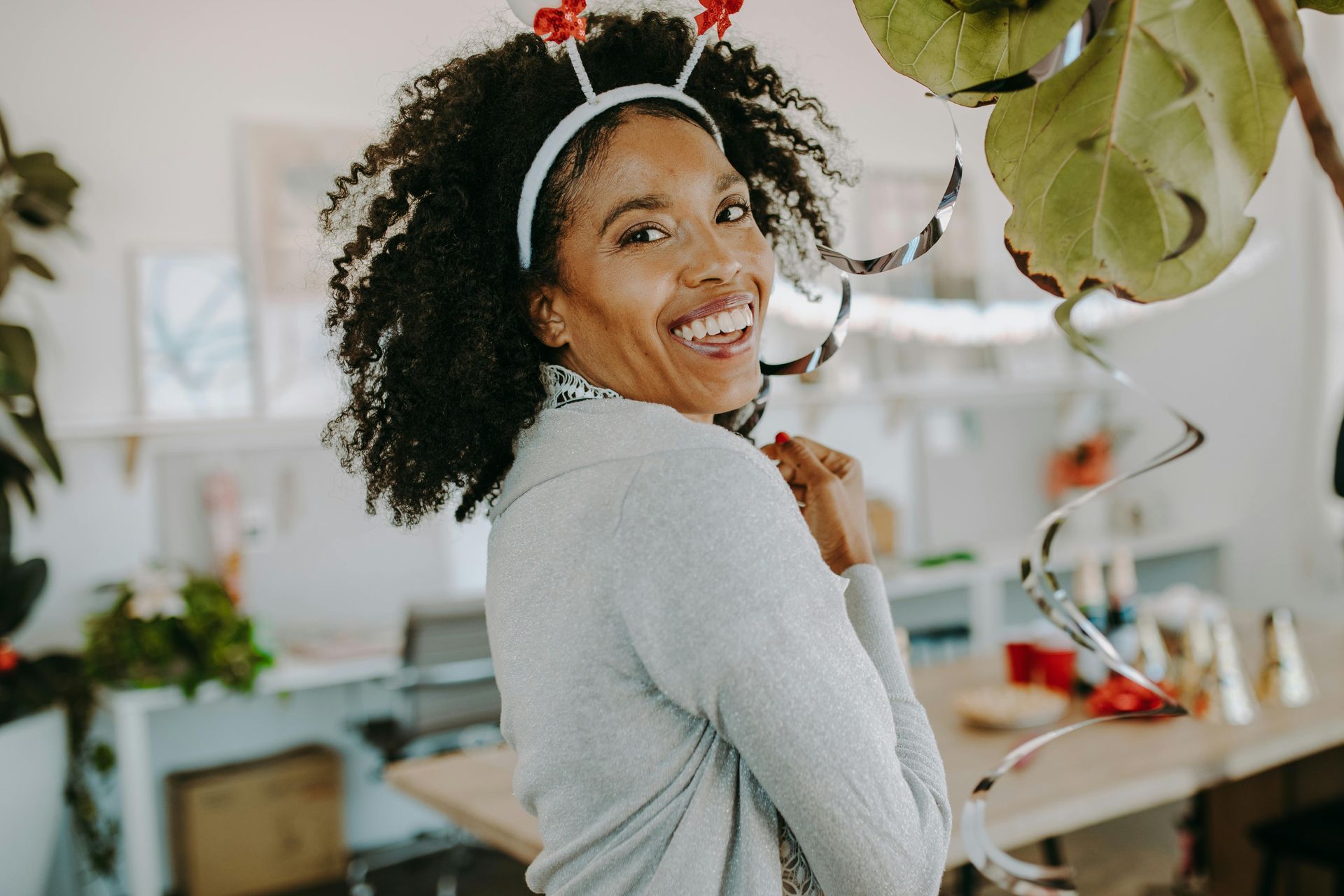 Woman smiling, wearing a reindeer antler headband. Indoors, festive decorations.