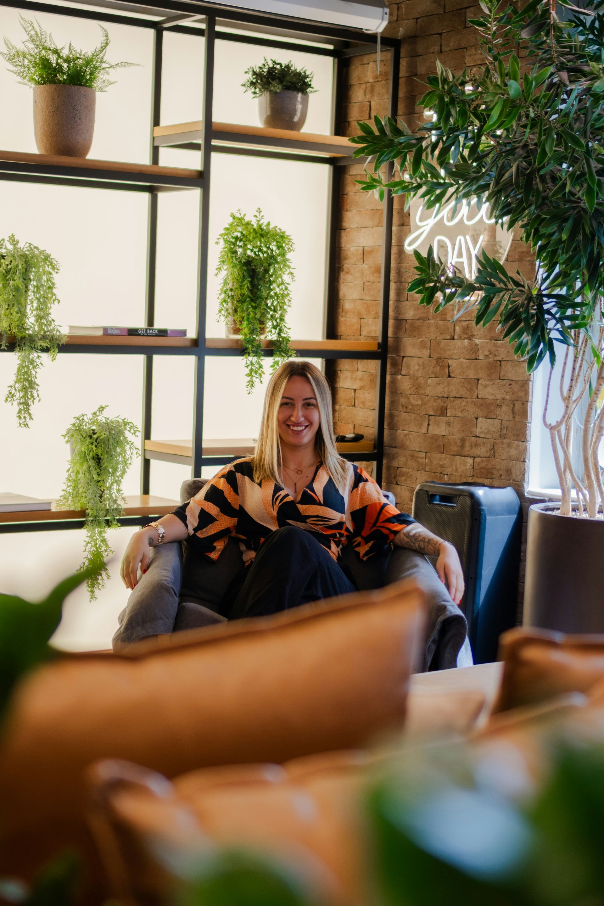 Woman sitting in chair, smiling, indoors. Plants on shelves and in pots behind. Brick wall, neon sign.