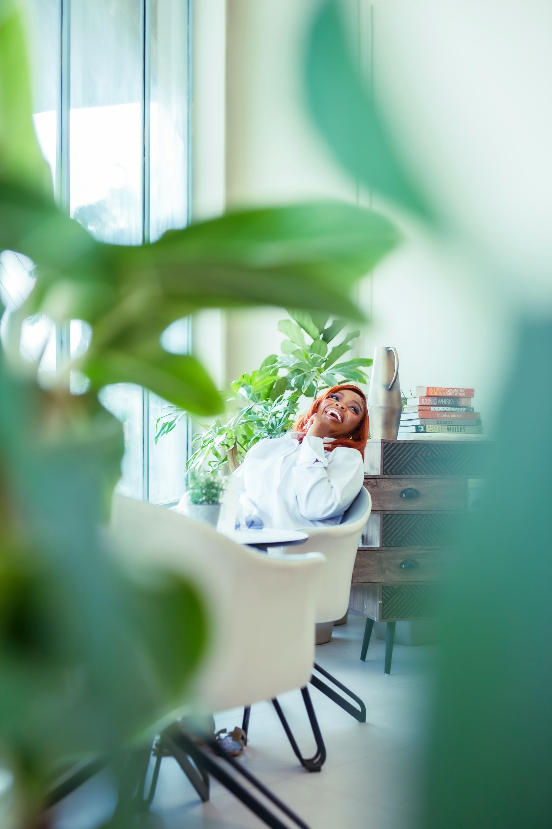 Woman relaxing in white chair by window, plants in foreground.