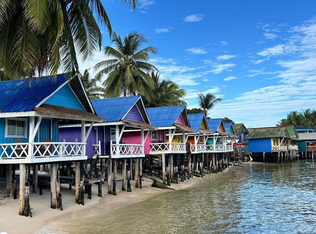 Colorful stilt houses with blue, purple, pink, and yellow roofs along a beach with palm trees under a bright sky.