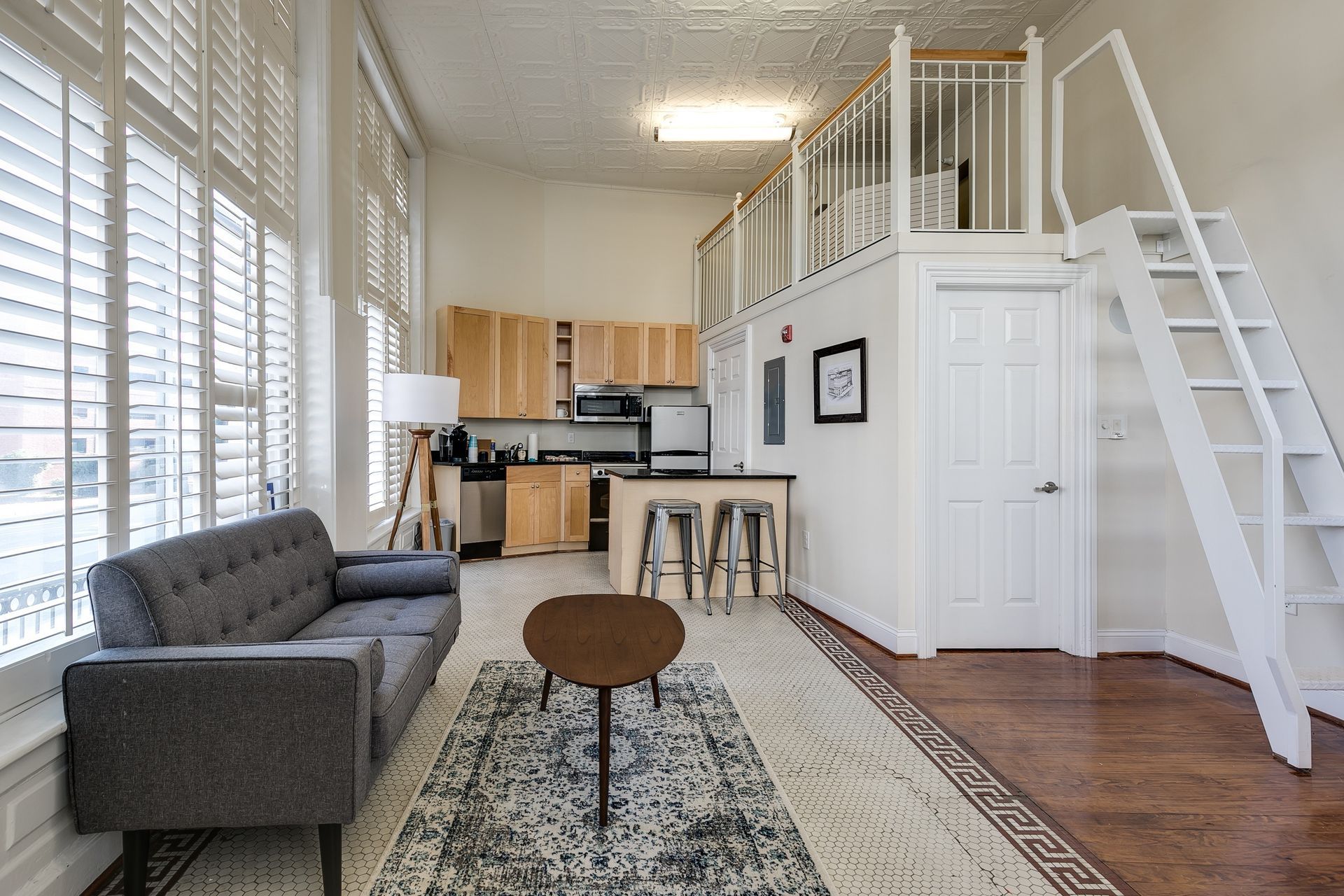 Loft apartment interior: gray sofa, kitchen, small table, rug, window shutters, and a lofted sleeping area.