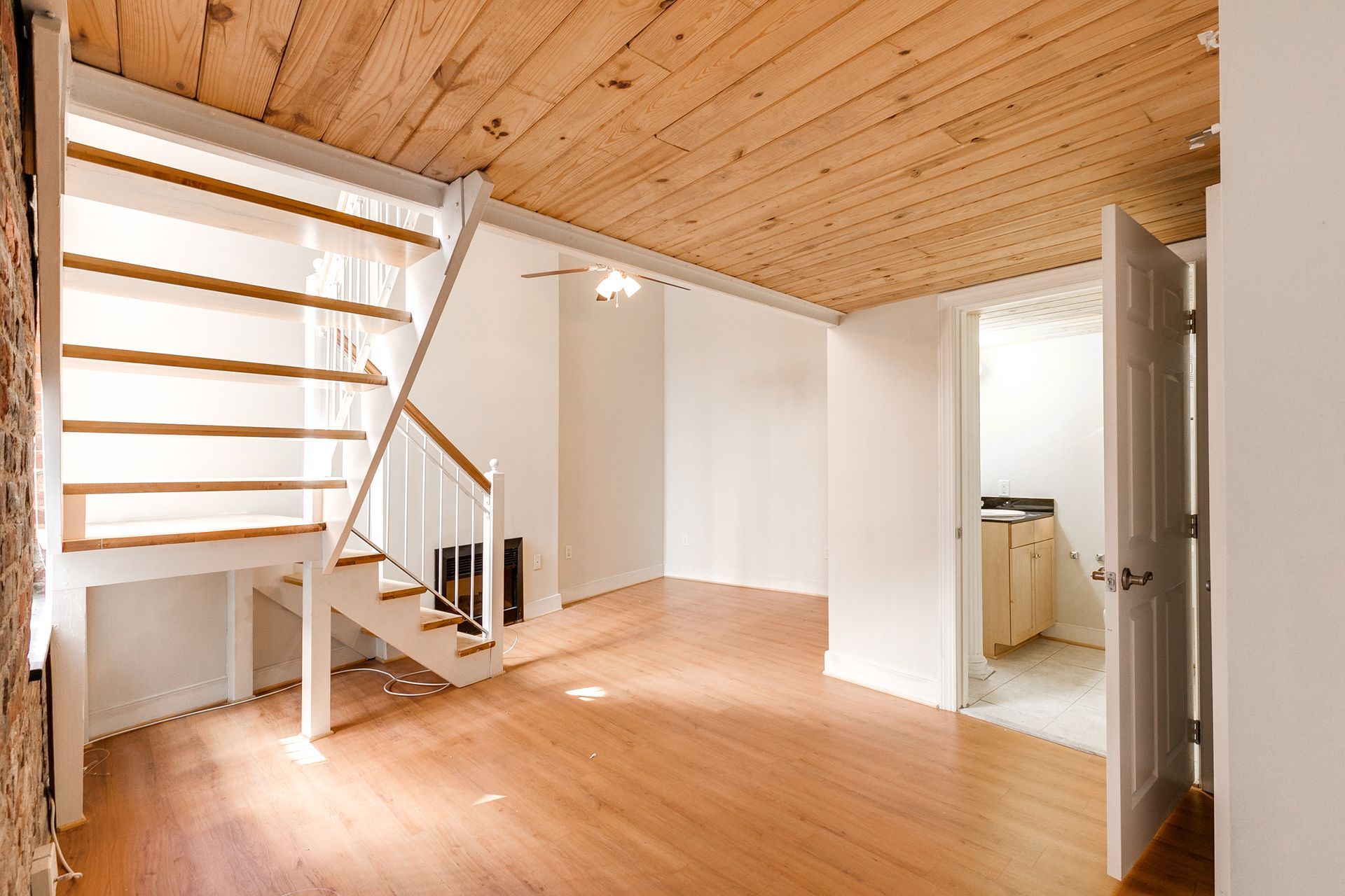 Interior with wooden floors, stairs, and ceiling. A doorway leads to a bathroom. Sunlight streams in.