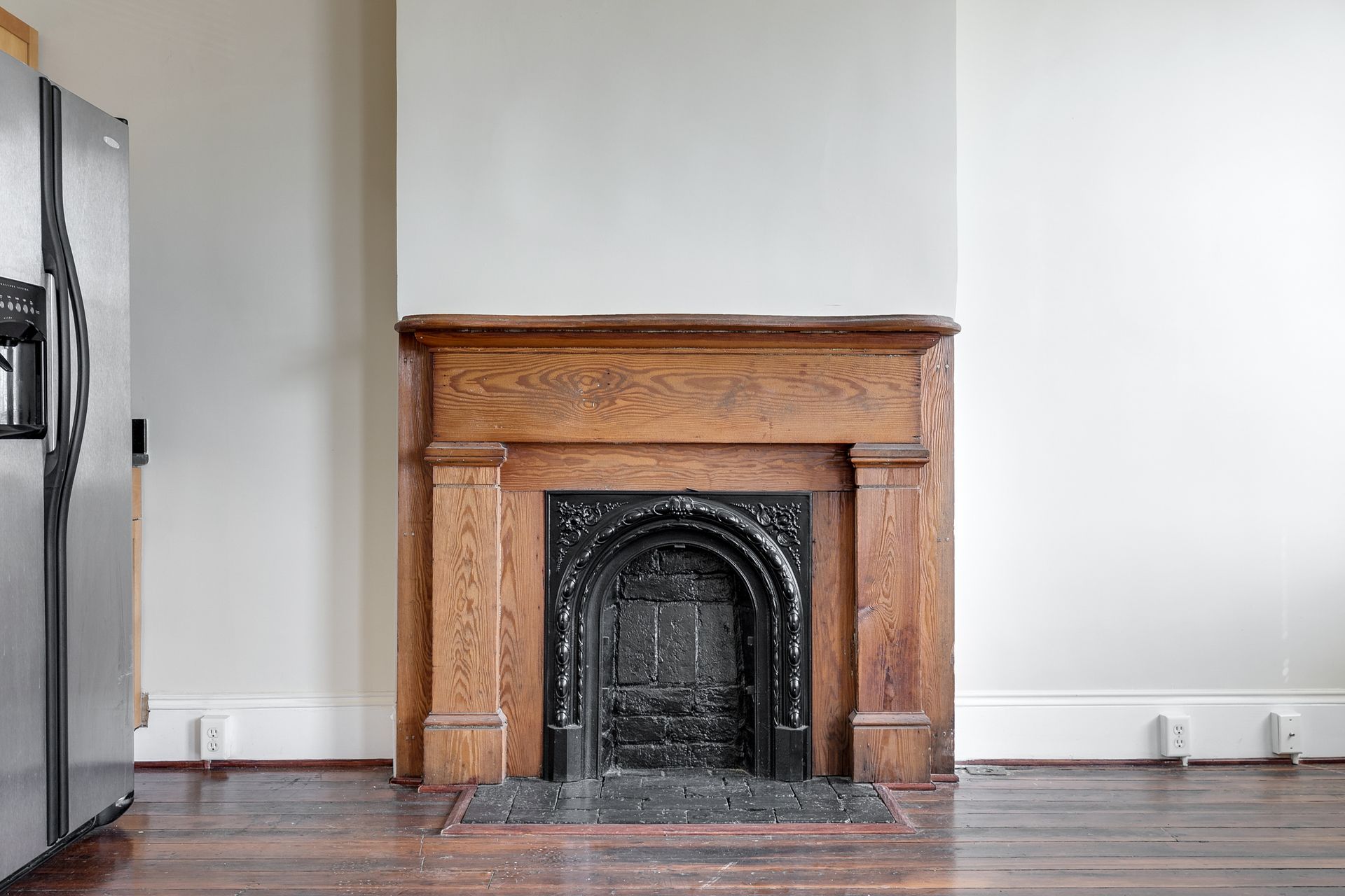 Wooden fireplace with a dark metal interior and decorative trim, set against a white wall in a room with hardwood floors.