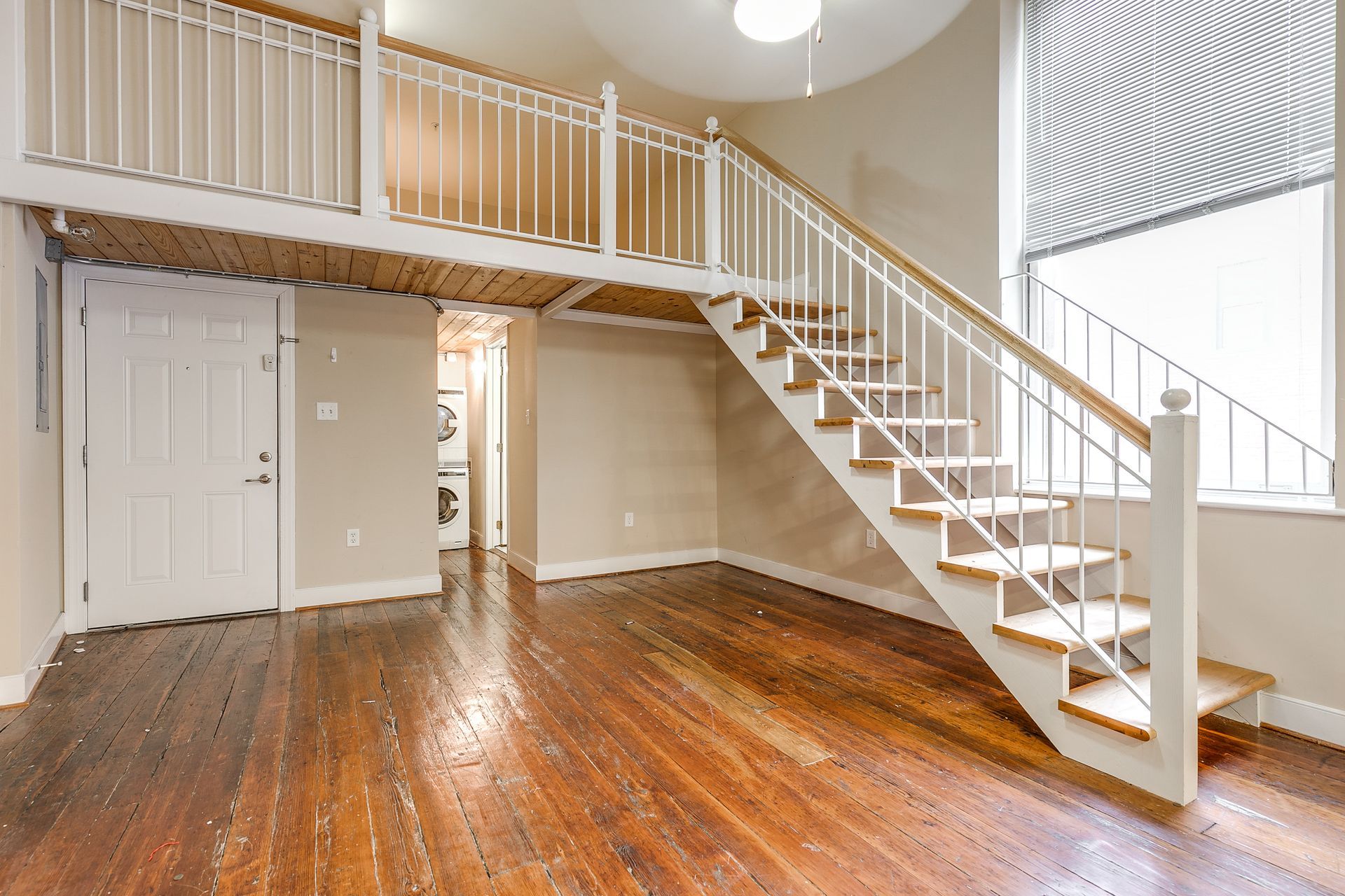 A bright loft interior with wooden floors, staircase leading to a mezzanine, and a large window.