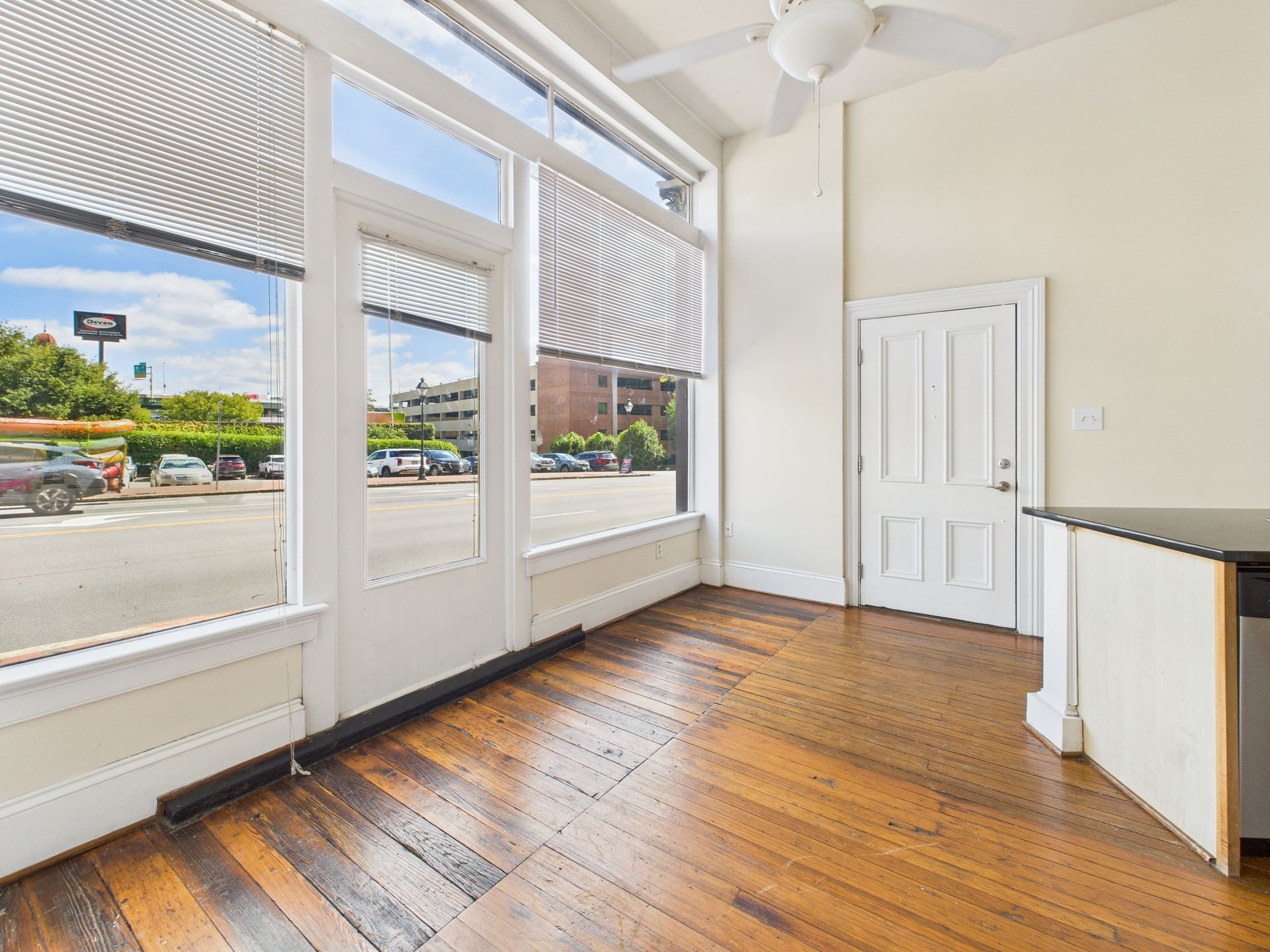Interior of a room with large windows overlooking a parking lot, hardwood floor, white door and ceiling fan.