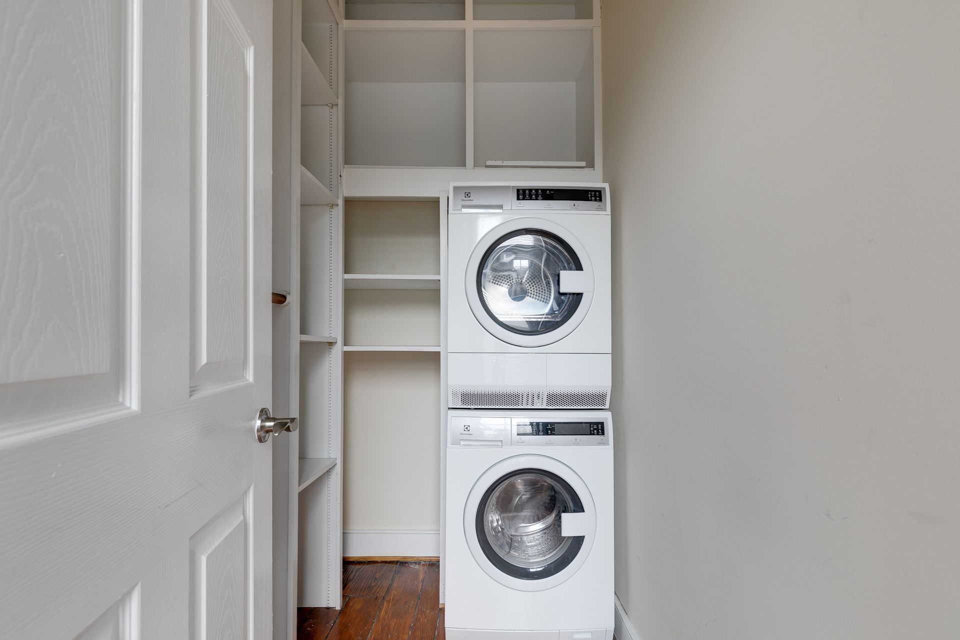 White stacked washer and dryer in a small laundry room, with built-in shelving and a closed white door.