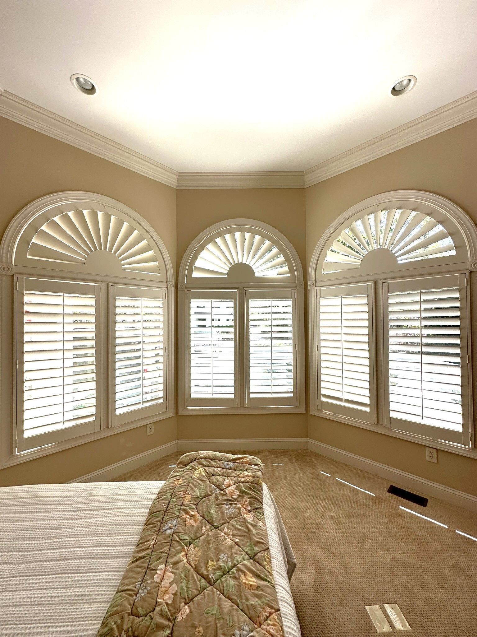 Bedroom with three arched windows, white shutters, beige walls, and light carpet. Bed with patterned bedspread.