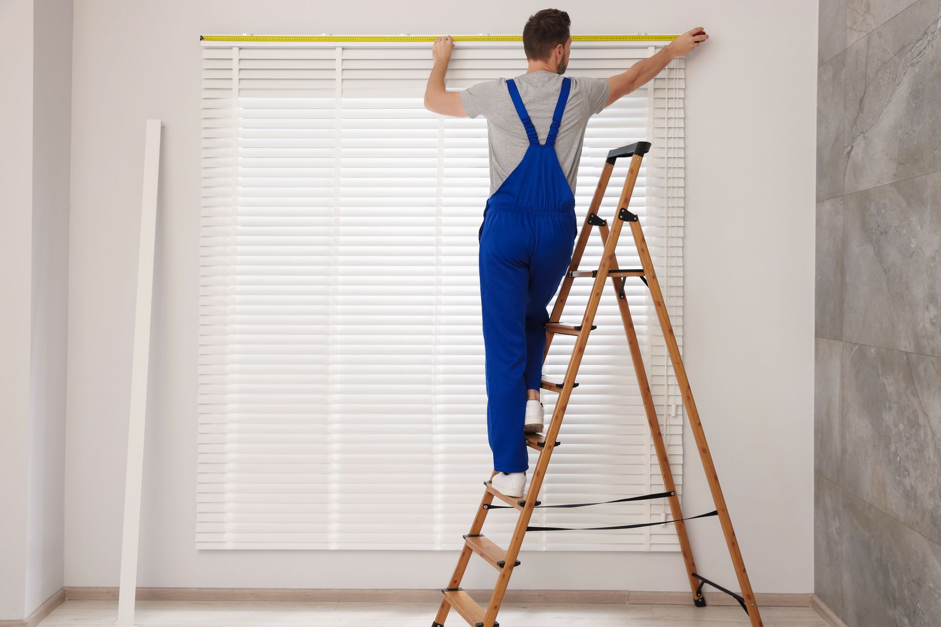 A man measuring window blinds from a ladder, preparing for custom window treatment installation.