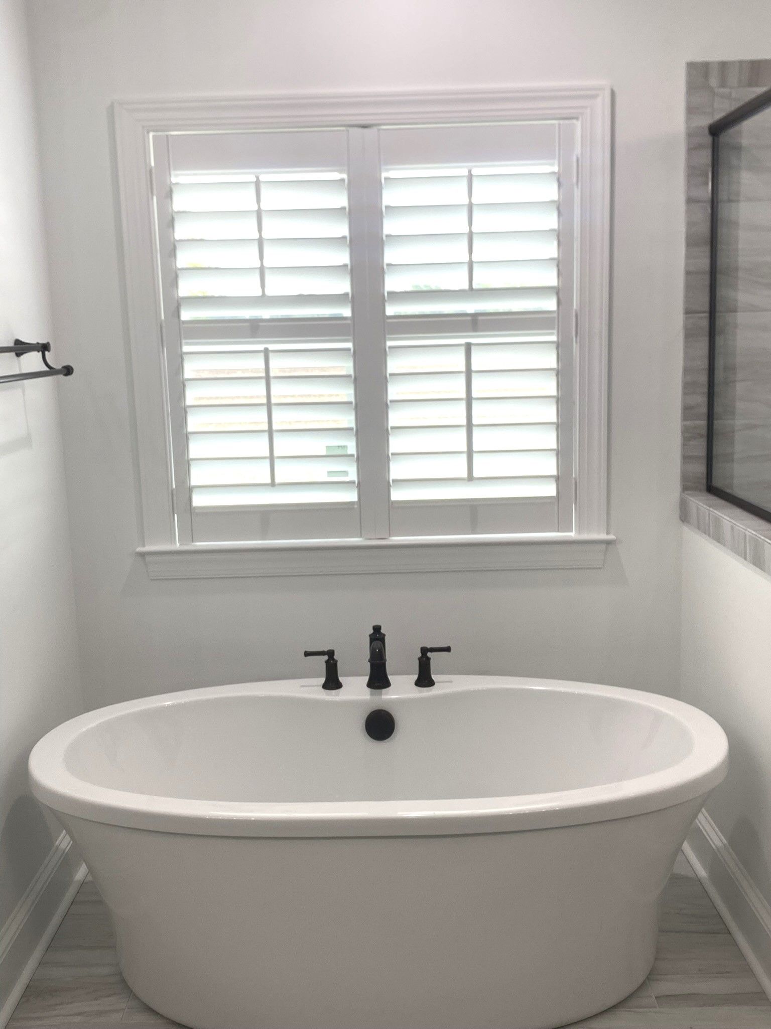 White soaking tub under a window with white shutters in a modern bathroom.