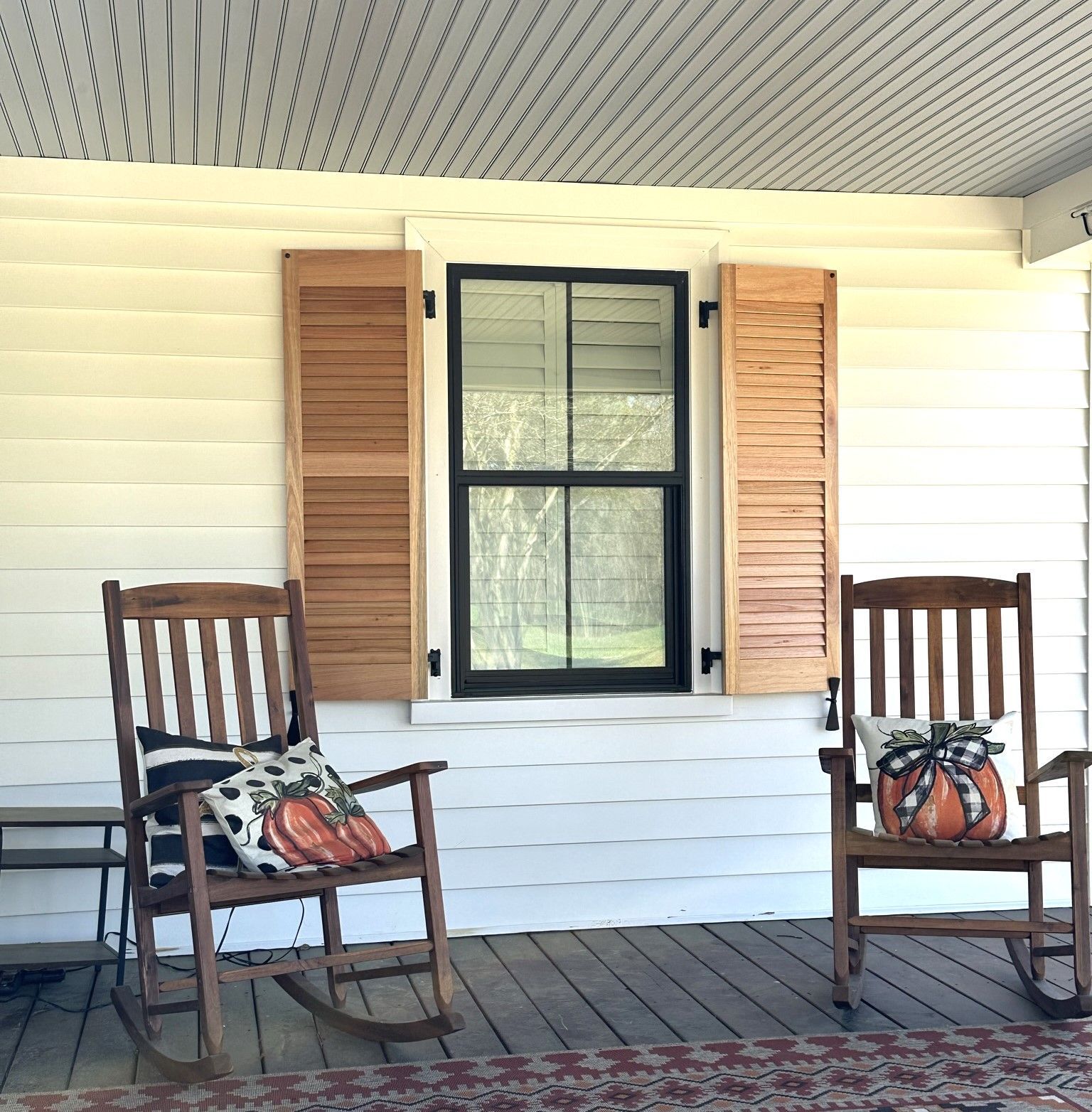 Two wooden rocking chairs on a porch with a window and pumpkin pillows.