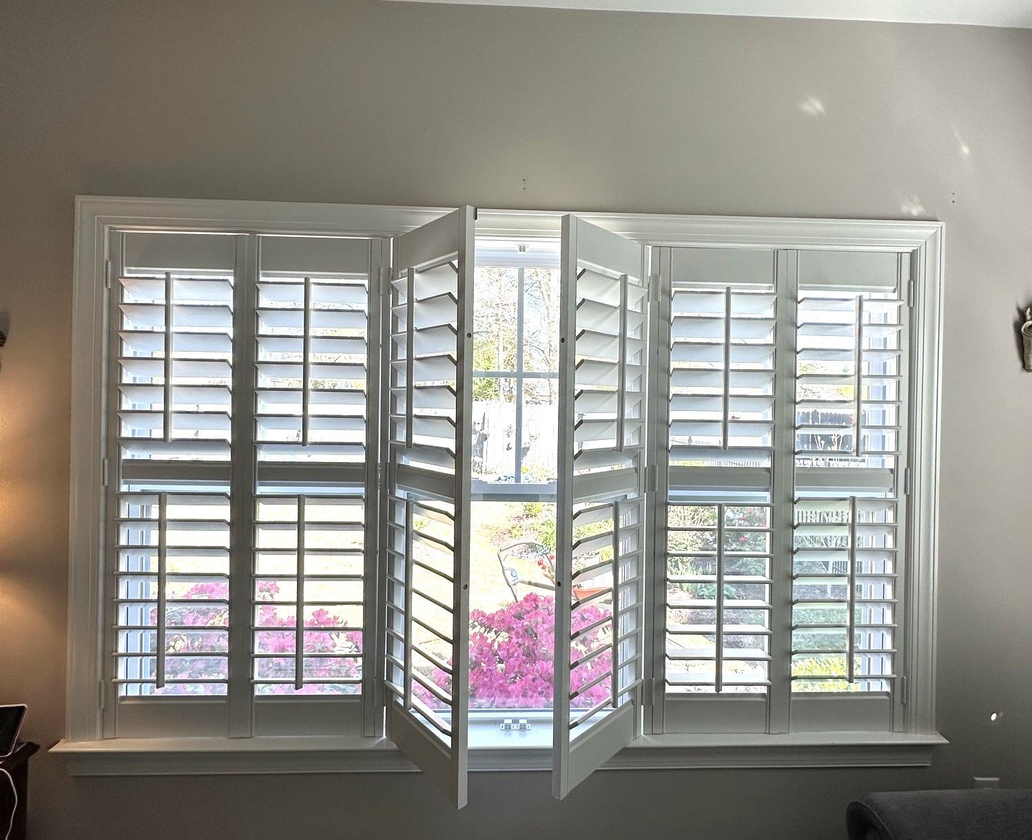White shutters on a window, with open center panels revealing a view of pink and green foliage.