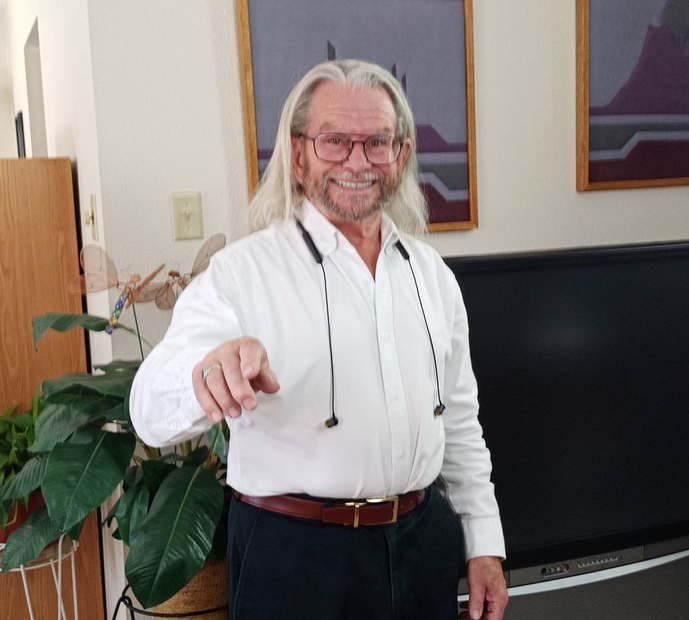 Older man with long white hair and glasses pointing, smiling. Indoors, near TV and plant.