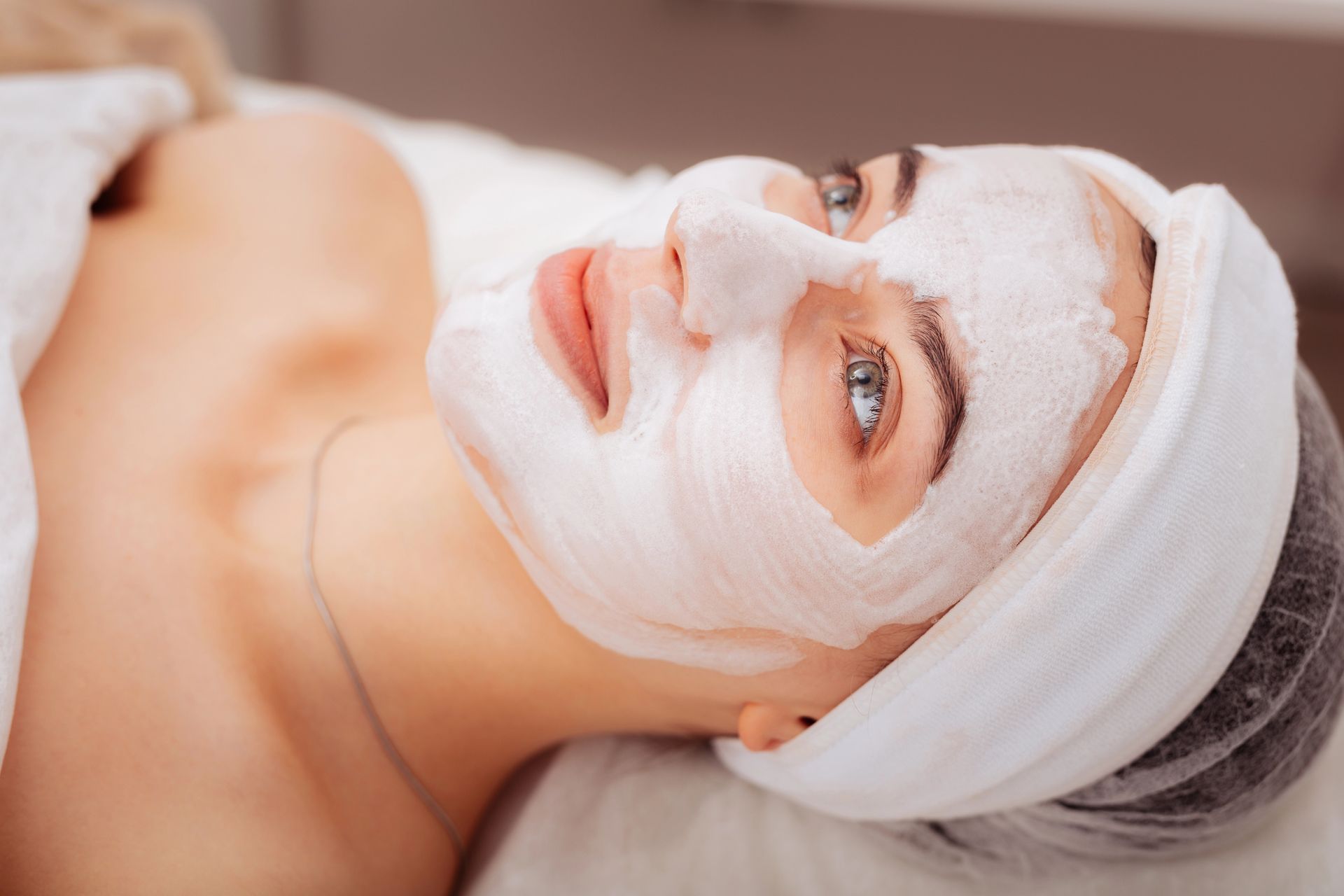 Donna con maschera facciale, sdraiata. Fascia per capelli, lenzuolo bianco, ambiente spa.