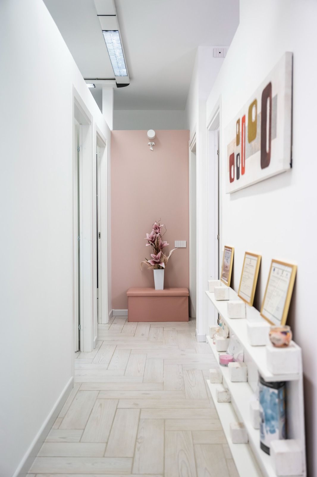 Stretto corridoio bianco con porte, una parete decorata in rosa, opere d'arte e uno scaffale con oggetti decorativi.