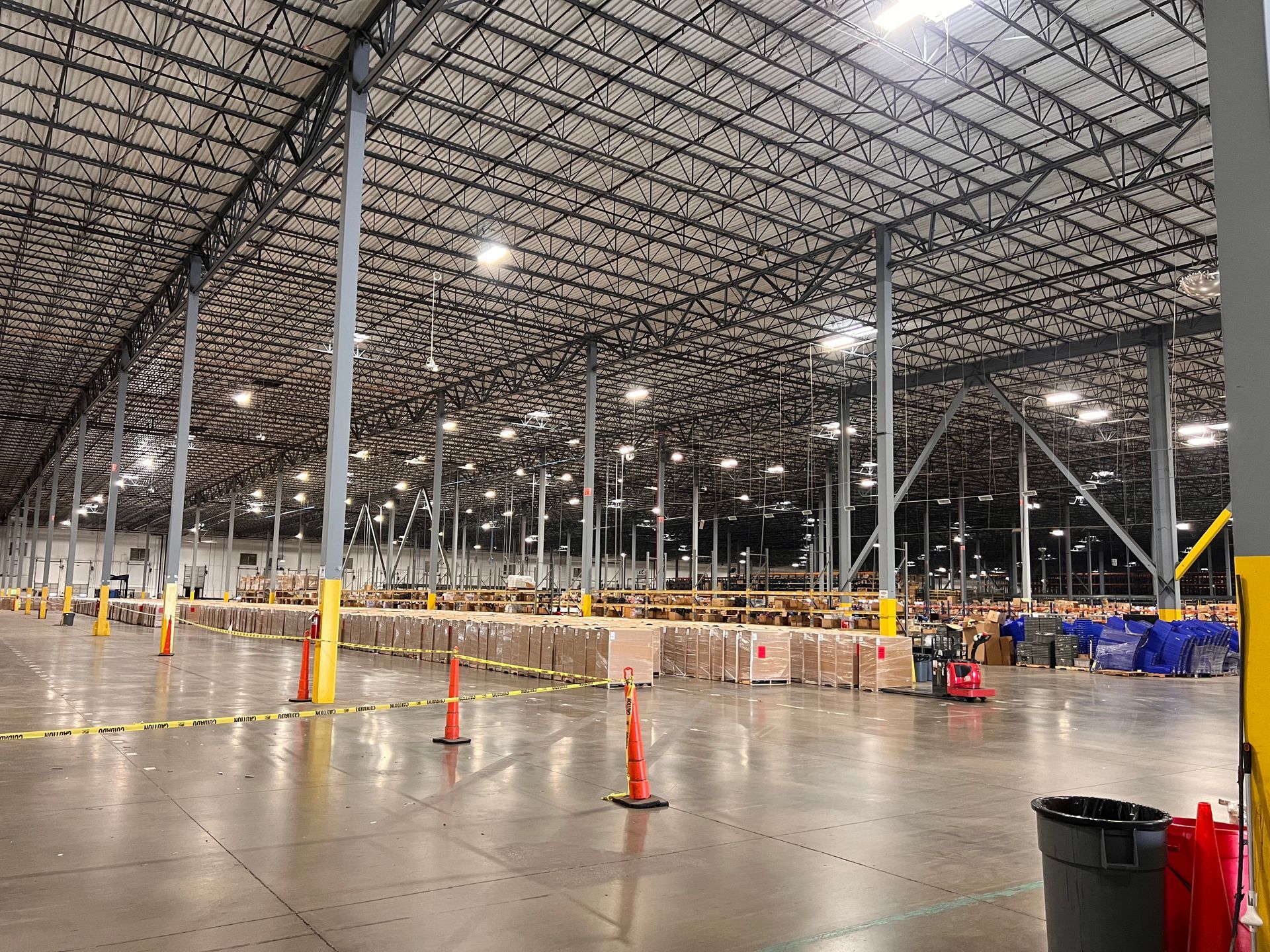 Spacious warehouse interior with high ceiling, orange cones, pallets, and a few workers near blue bins
