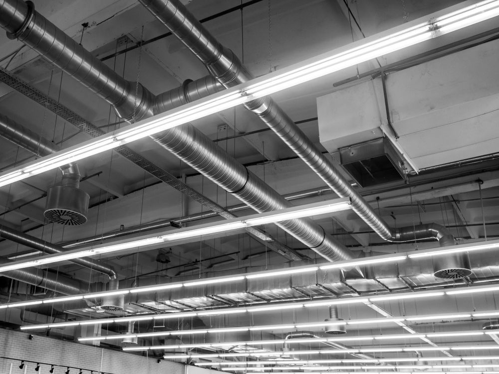 Black and white photo of a high industrial ceiling with exposed pipes, ventilation, and bright fluorescent lights.