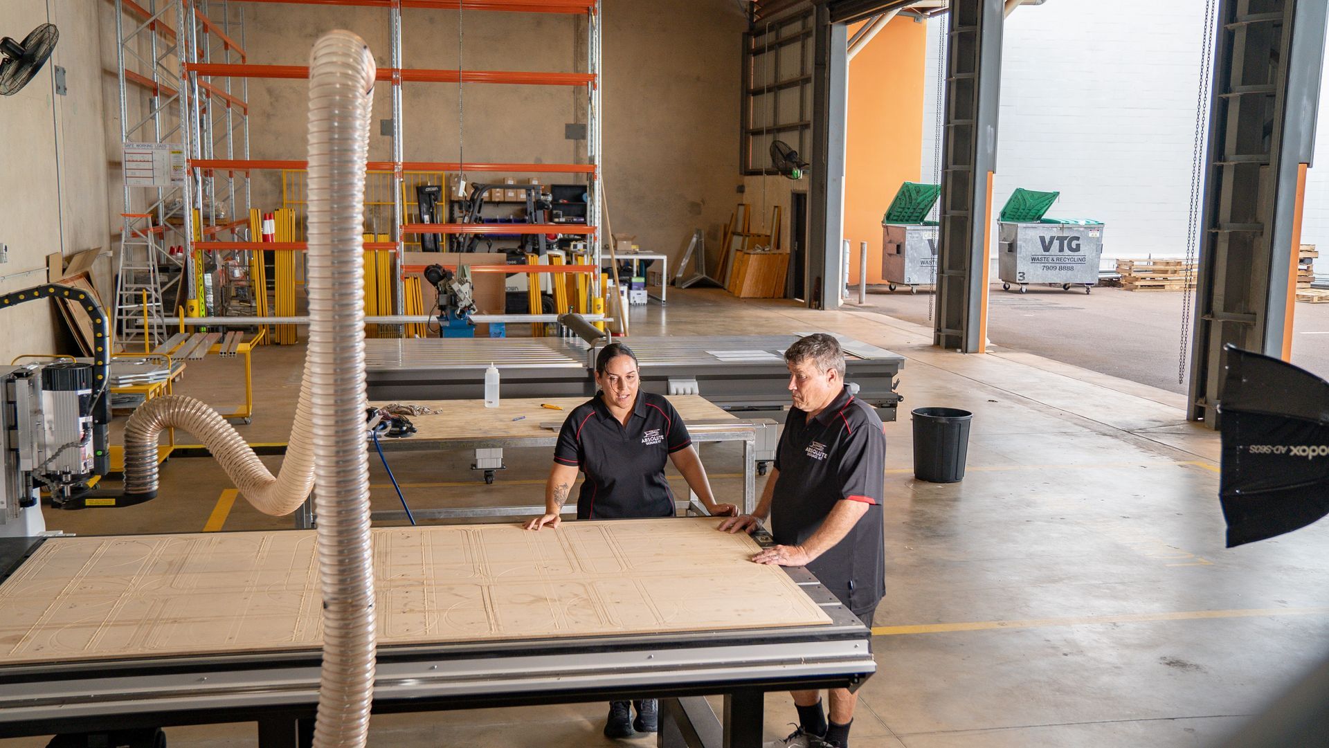 Two people in workshop, looking at a CNC machine cutting wood. Building in background.