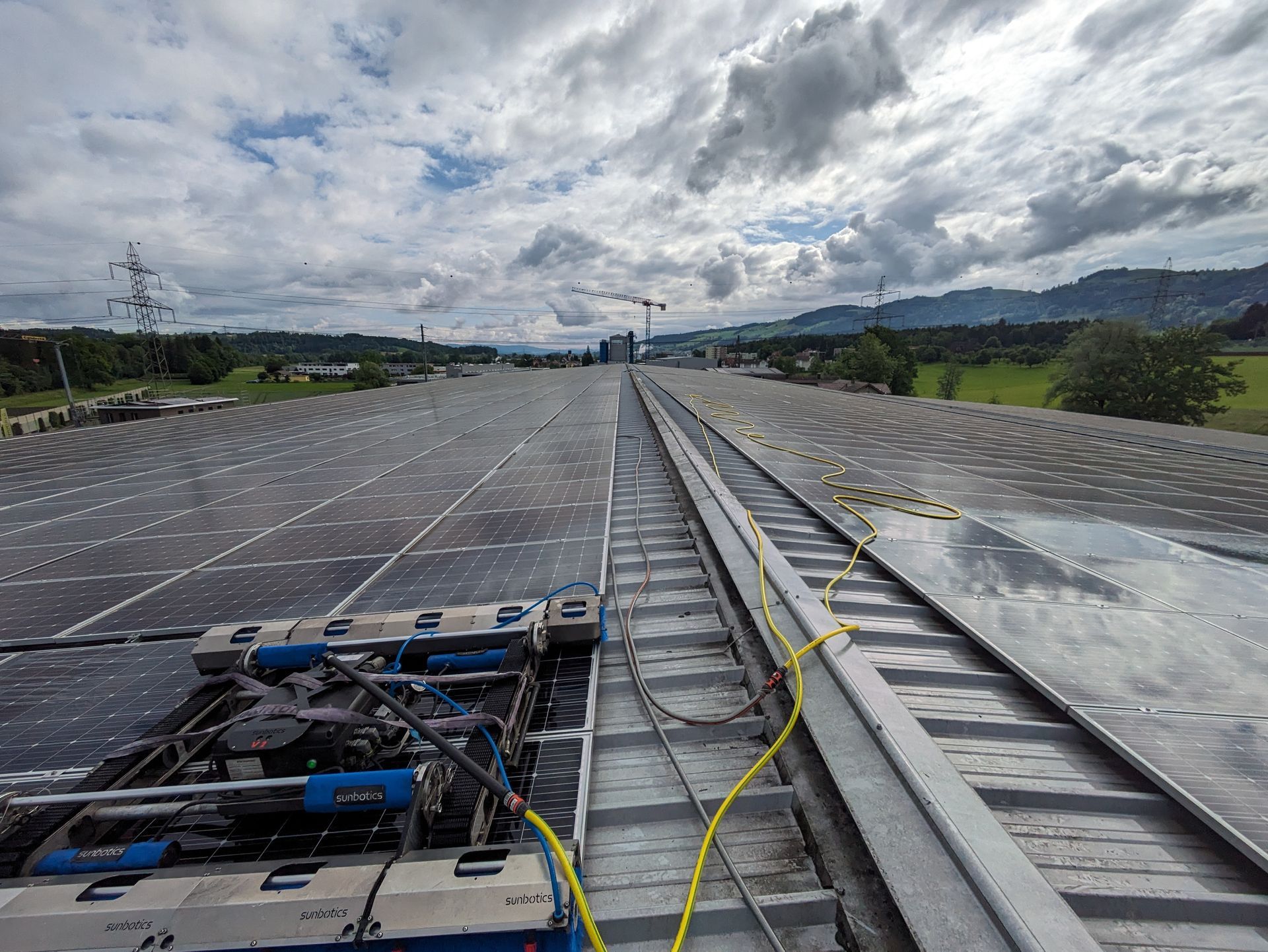 Solaranlage auf einem Dach mit einem Reinigungsroboter, bewölktem Himmel und grünen Hügeln in der Ferne.