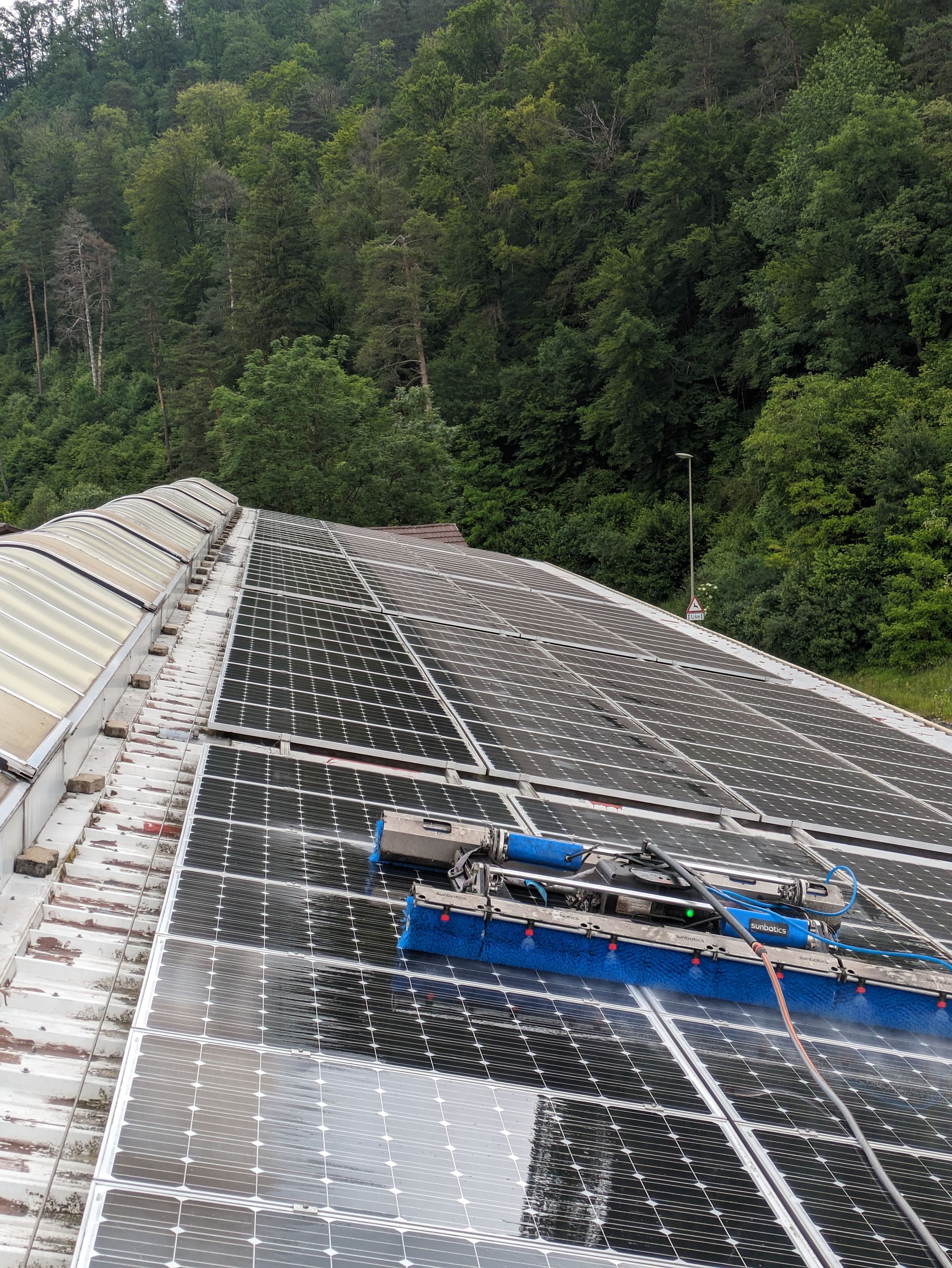 Solarpaneele auf einem Dach werden von einem automatischen Bürstensystem gereinigt; im Hintergrund sind Bäume zu sehen.