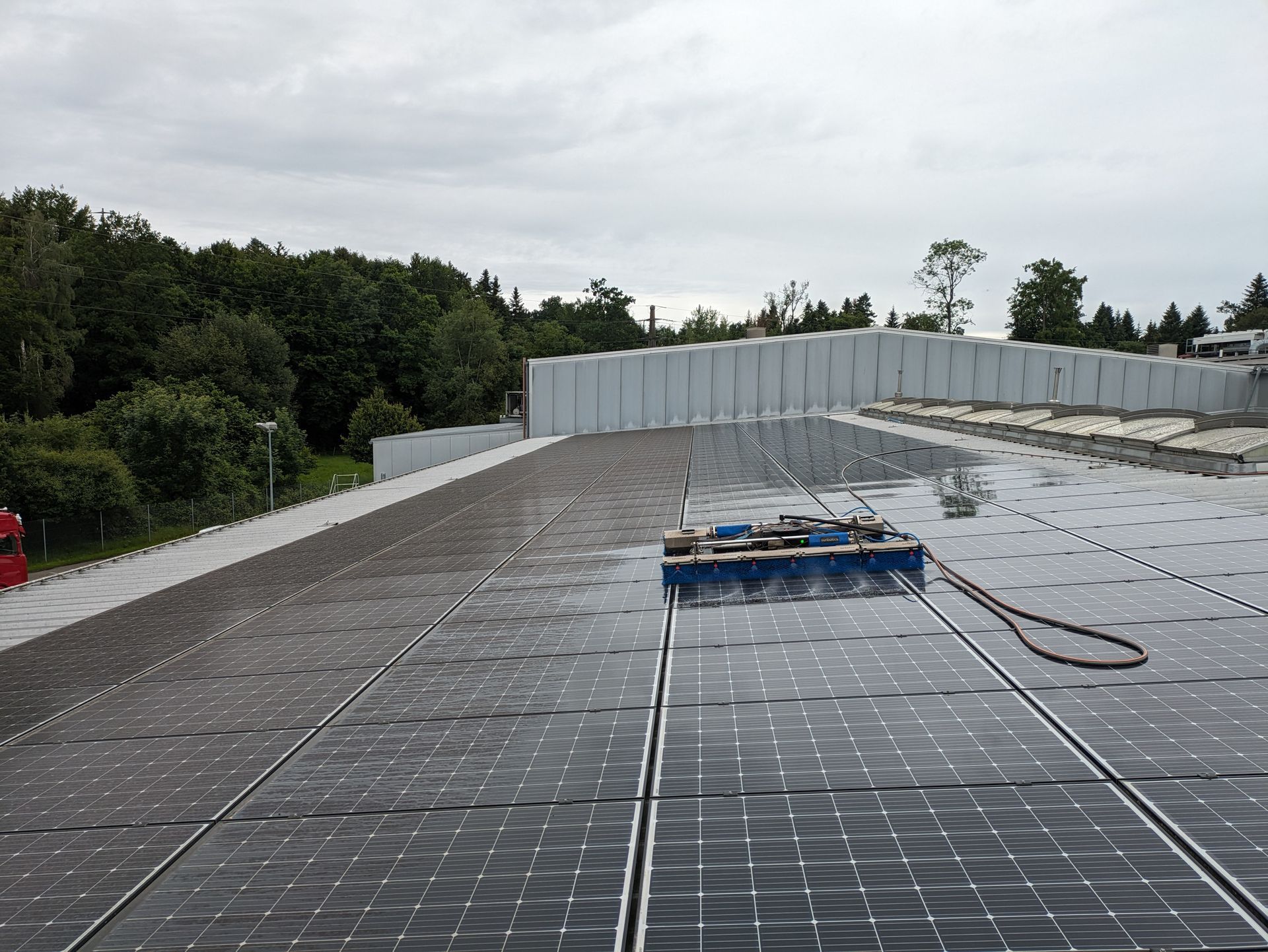 Auf einem Gebäudedach werden Solarpaneele von einem Reinigungsroboter gesäubert, im Hintergrund sind Bäume zu sehen.