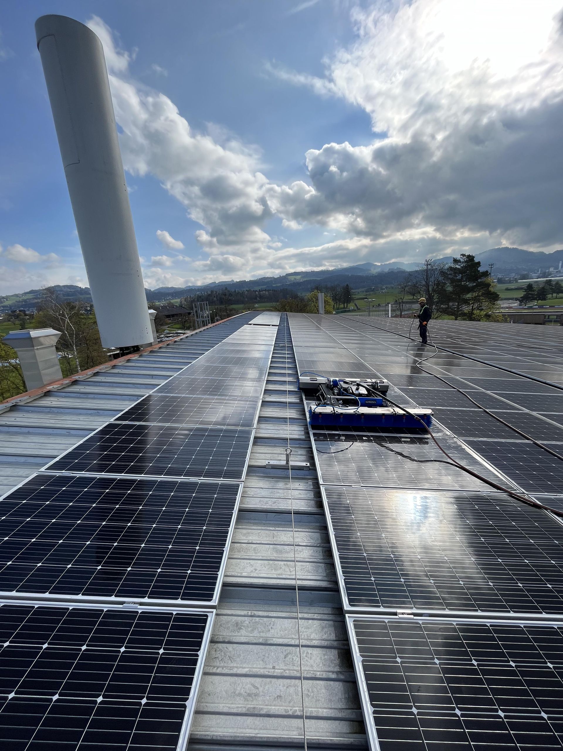 Auf einem Dach werden Solarpaneele gereinigt; im Hintergrund sind Berge und ein bewölkter Himmel zu sehen.