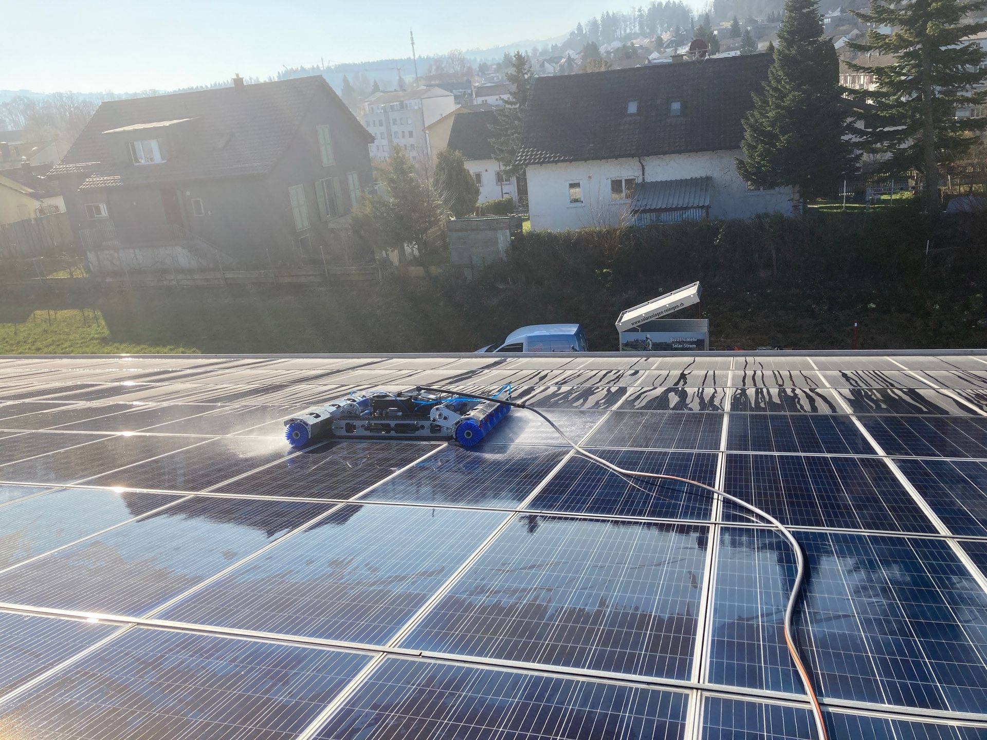 Auf einem Dach werden Solarpaneele von einem blauen Robotergerät gereinigt; im Hintergrund ist eine Stadt zu sehen.
