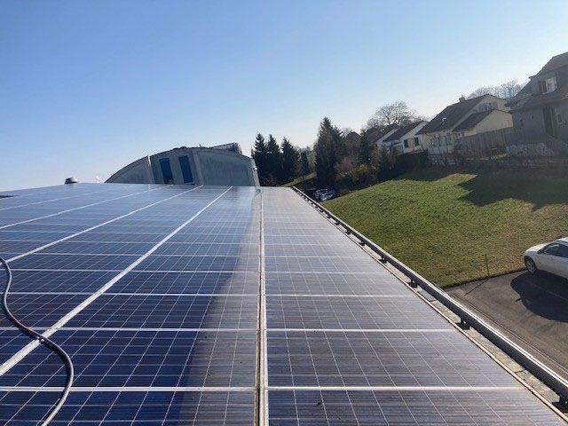 Solarmodule auf einem Dach, sonniger Tag. Die Module bedecken den größten Teil des Daches; im Hintergrund Häuser und Hügel.