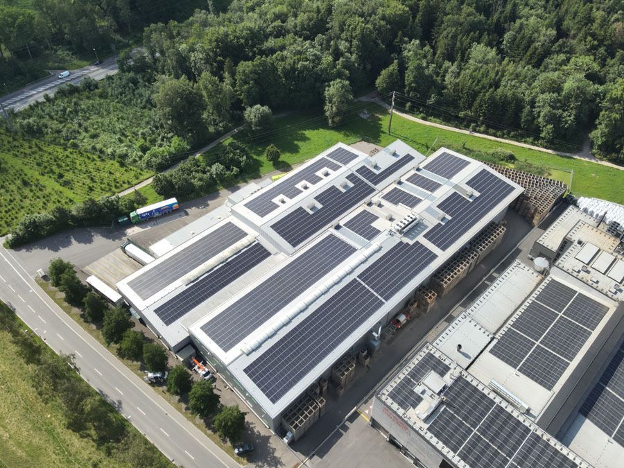 Luftaufnahme eines mit Solarpaneelen bedeckten Fabrikdachs, umgeben von Bäumen und einer Strasse.