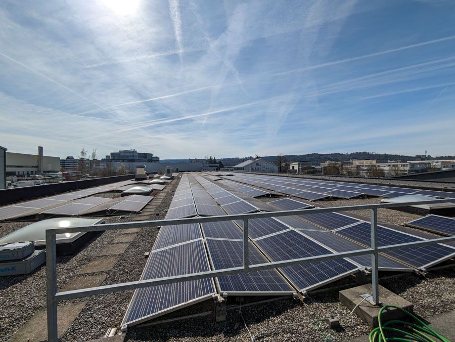 Solarmodule auf einem Flachdach vor blauem Himmel, von einem Metallgeländer aus betrachtet.