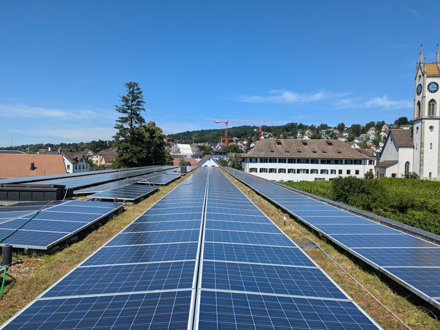 An einem sonnigen Tag bedecken Solarmodule ein Dach, im Hintergrund sind eine Kirche und Häuser zu sehen.