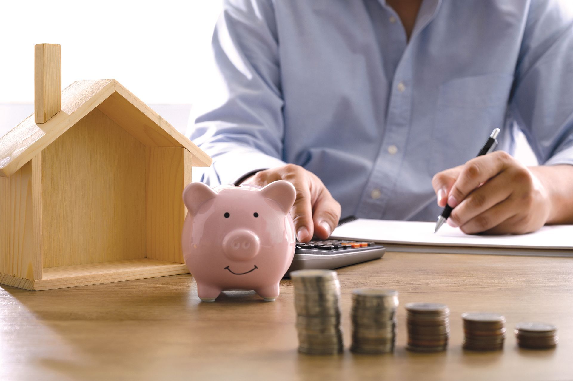 Person calculating finances at a desk with a piggy bank, wooden house, and decreasing coin stacks.