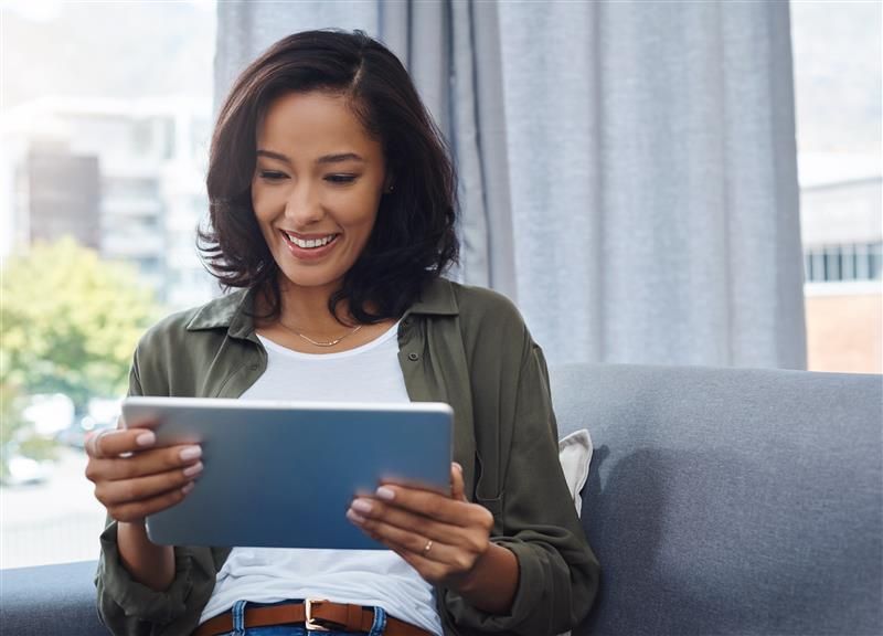 Woman smiling while holding tablet, sitting on couch near a window.
